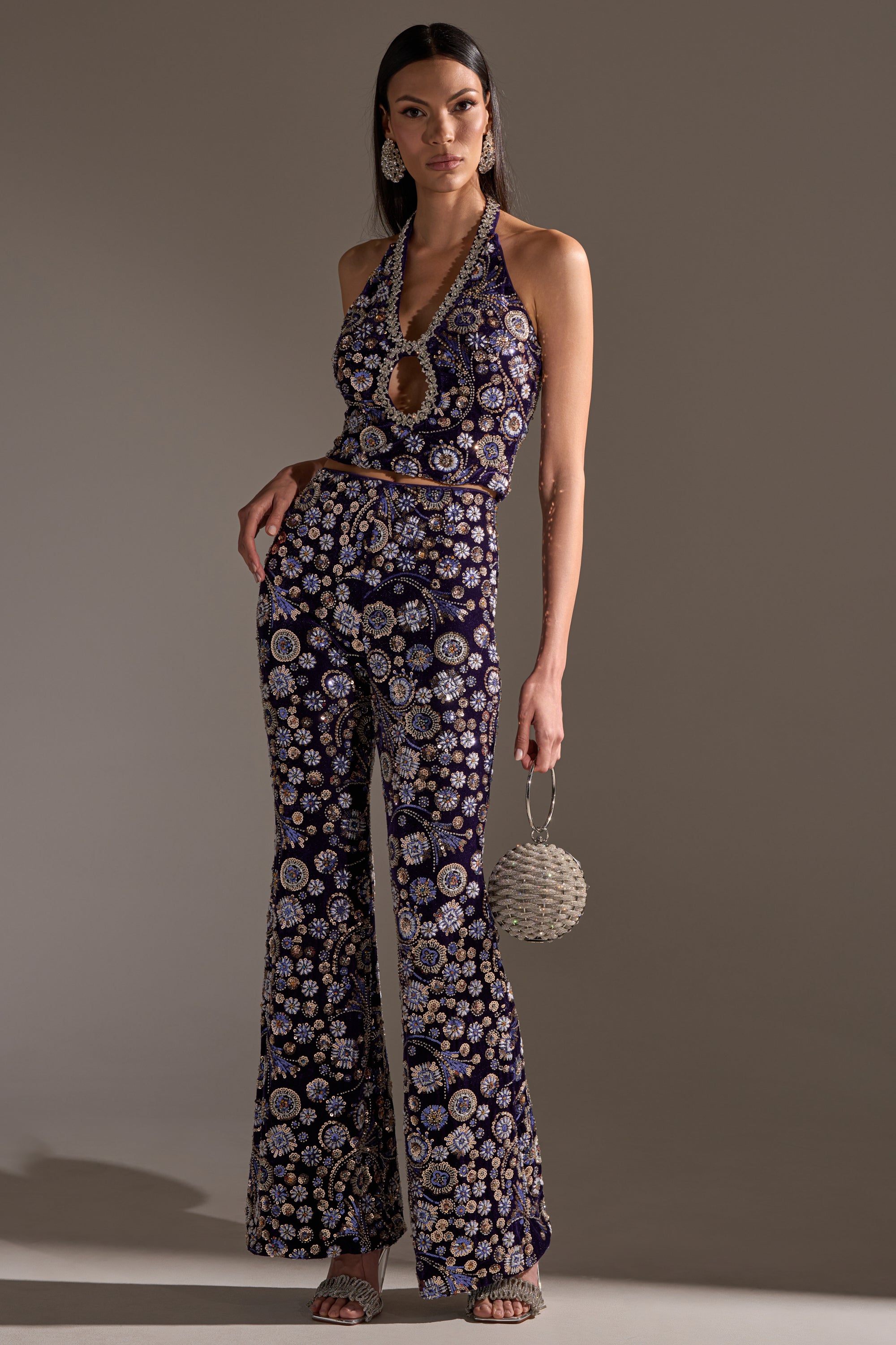 A woman showcases the QUEEN OF EVERYTHING RHINESTONE TROUSER, a sleeveless, halter-neck jumpsuit with keyhole cutout and flared legs, adorned with floral patterns and subtle rhinestone details. She accessorizes with a beaded bag, statement earrings, and strappy heels.