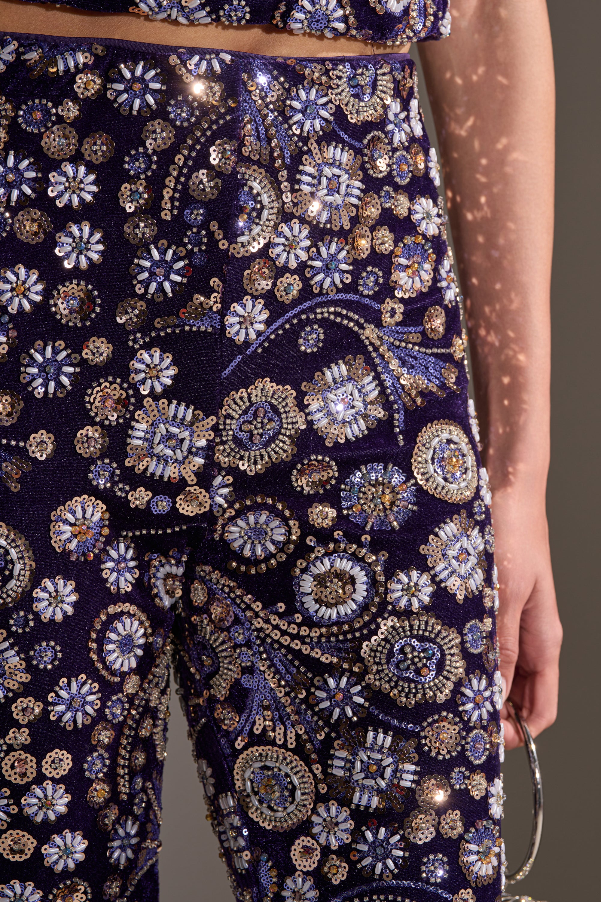 Close-up of a person wearing the QUEEN OF EVERYTHING RHINESTONE TROUSER, high-rise pants covered in colorful floral beadwork and sequins for a sparkling, ornate look. The matching top and metallic handle complete the bold ensemble.
