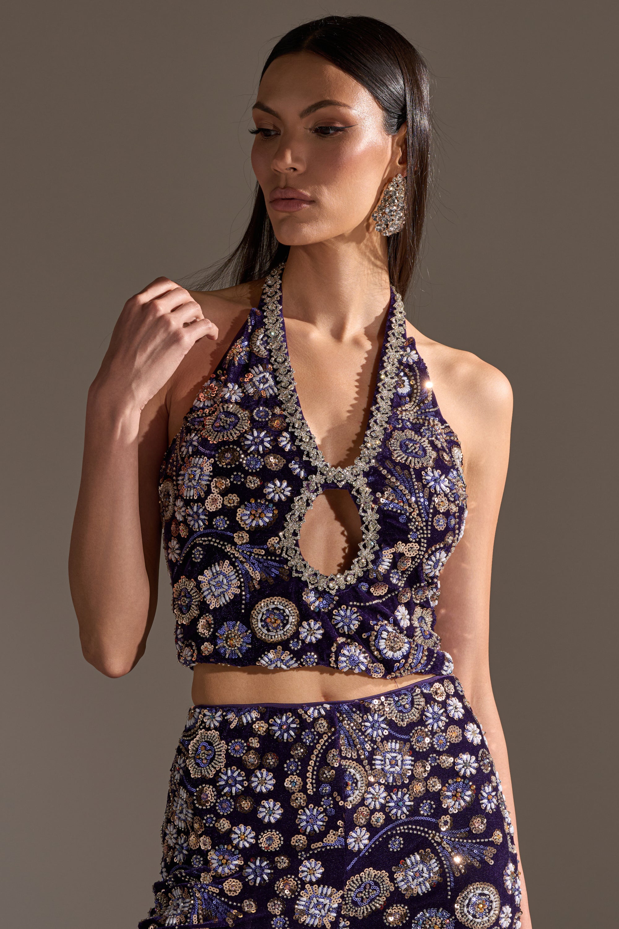 A woman wears the QUEEN OF EVERYTHING RHINESTONE HALTER BLOUSE, a deep purple cropped halter top with silver and gold rhinestone embroidery, paired with a matching skirt. She has long dark hair, statement earrings, and a neutral expression.