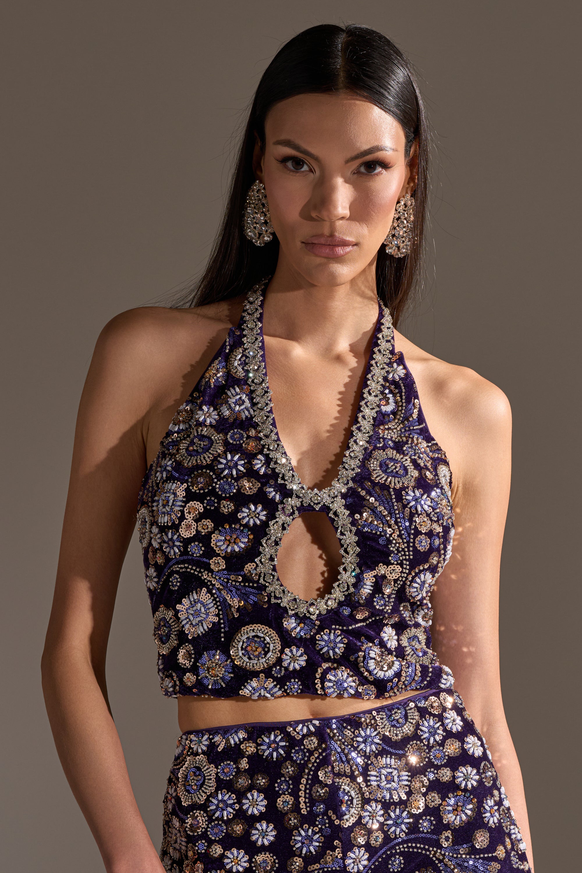 A woman with long dark hair wears the QUEEN OF EVERYTHING RHINESTONE HALTER BLOUSE, featuring intricate gold and silver floral embroidery. She pairs it with a matching skirt, ornate earrings, and looks confidently at the camera.