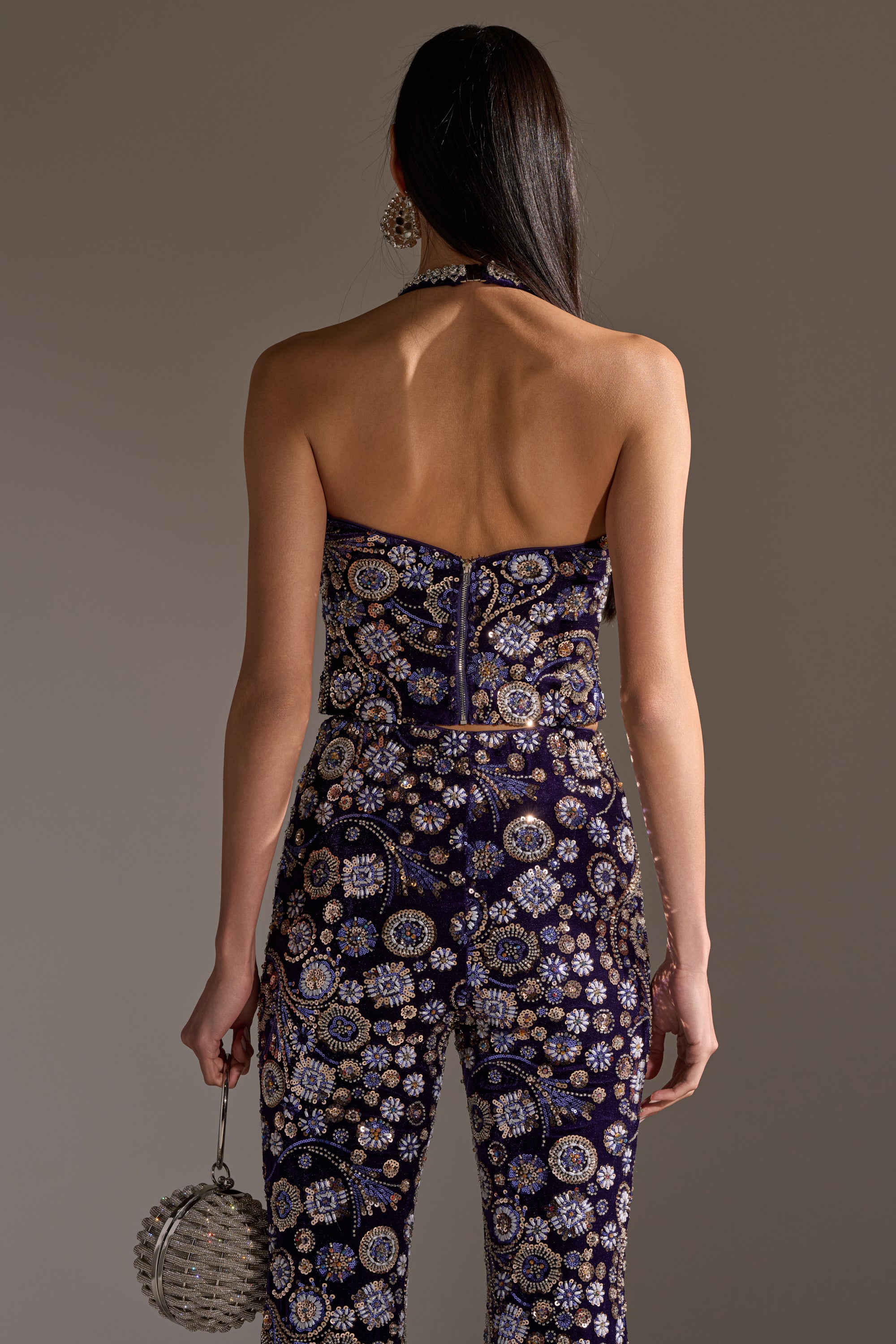 A woman with long dark hair stands facing away, wearing the QUEEN OF EVERYTHING RHINESTONE HALTER BLOUSE, featuring intricate beadwork and floral patterns. She holds a round silver beaded clutch in her left hand.