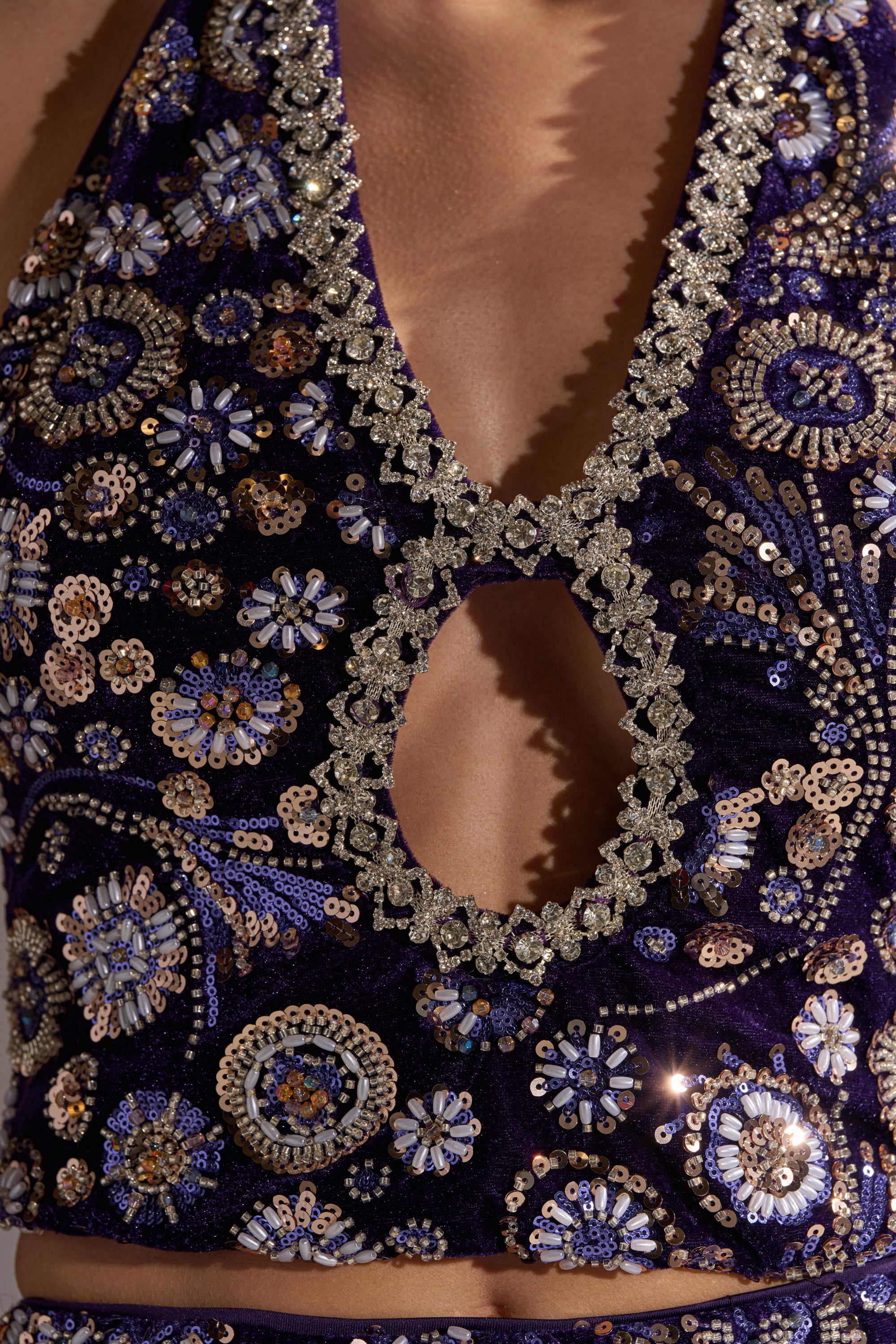 A close-up of the QUEEN OF EVERYTHING RHINESTONE HALTER BLOUSE, a deep purple velvet top featuring intricate floral sequin patterns, sparkling beadwork, a cropped fit, plunging neckline with a circular cutout, and shimmering embellishments.