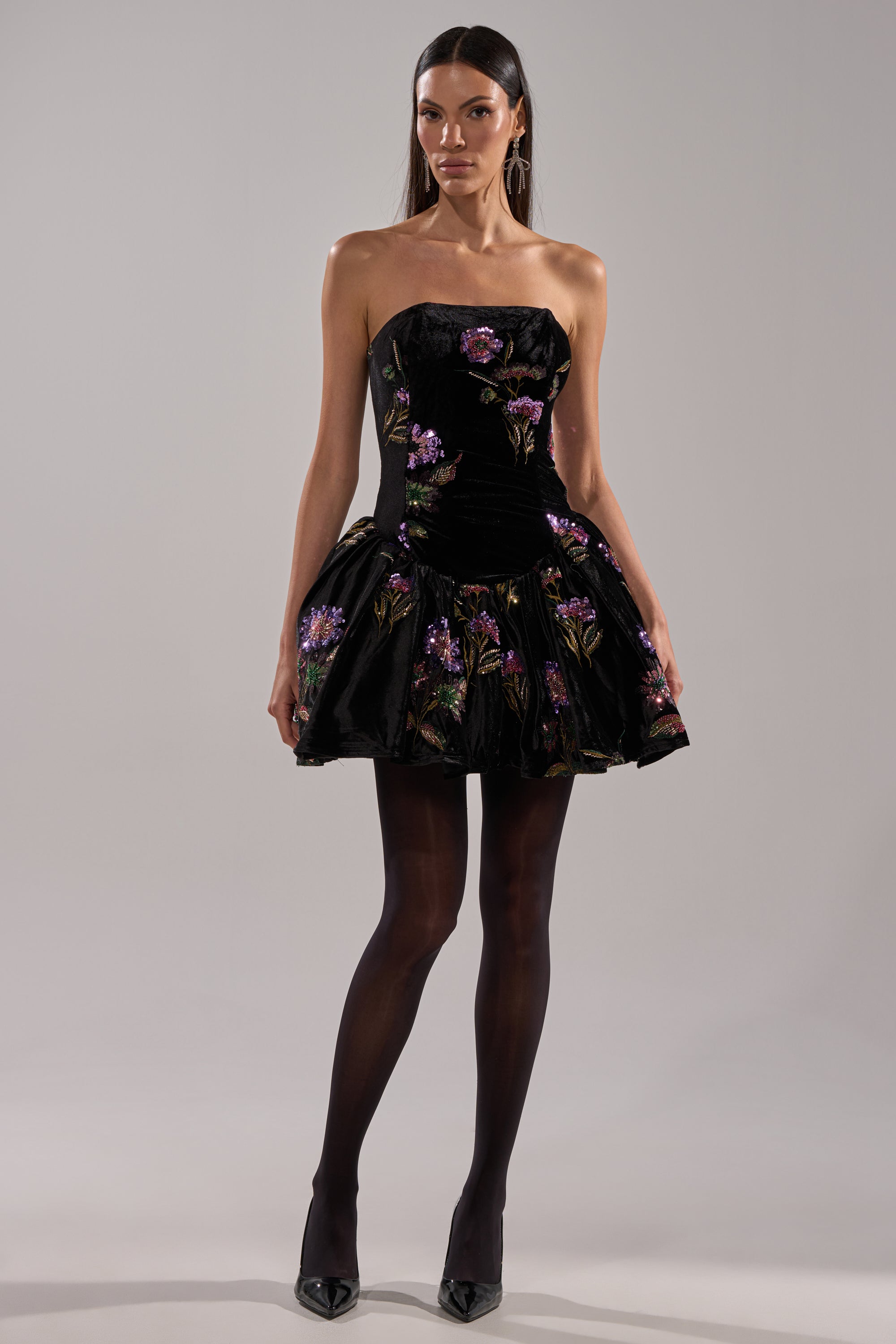 A woman faces forward in the LATE NIGHT GARDEN PARTY VELVET MINI DRESS, a strapless black mini with floral embroidery and a bubble skirt. She pairs it with black tights, heels, straight dark hair, and stands before a plain gray background.