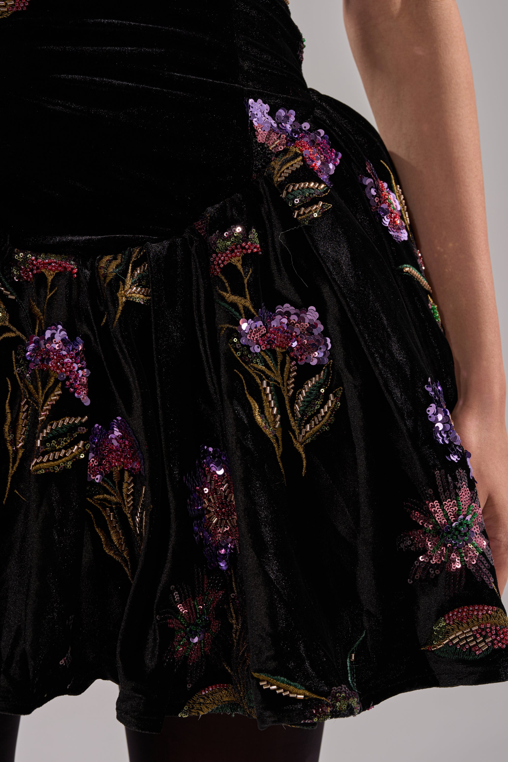 A close-up of a person wearing the LATE NIGHT GARDEN PARTY VELVET MINI DRESS, featuring black velvet with colorful embroidered and sequined floral patterns, their arm resting at their side.