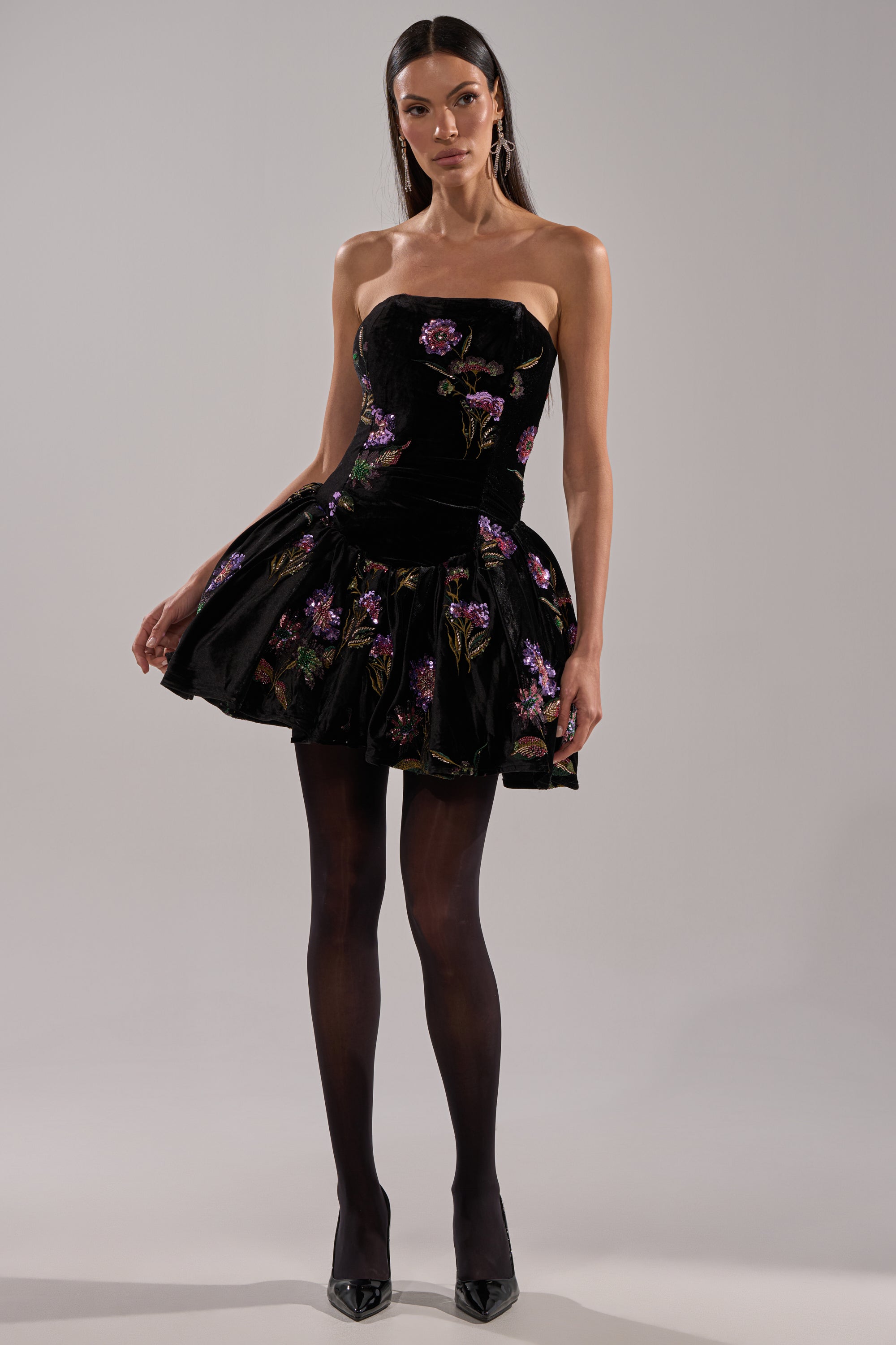 A woman wears the LATE NIGHT GARDEN PARTY VELVET MINI DRESS—a strapless black velvet mini dress with purple floral embroidery—paired with black sheer tights and pointed heels, standing against a plain backdrop.