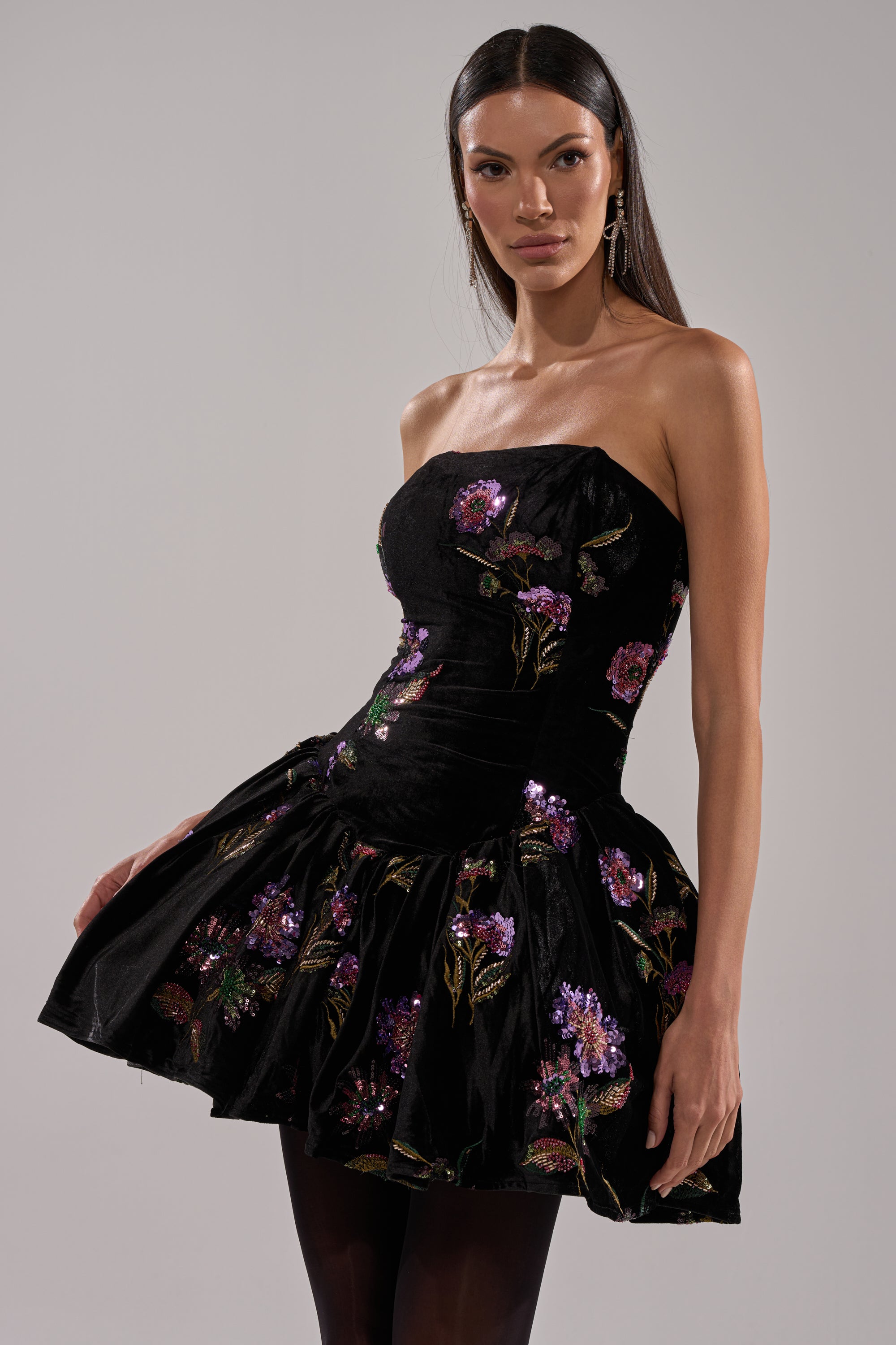 A woman models the LATE NIGHT GARDEN PARTY VELVET MINI DRESS, a strapless black mini dress with a flared bubble skirt and colorful floral embroidery, standing against a plain light background.