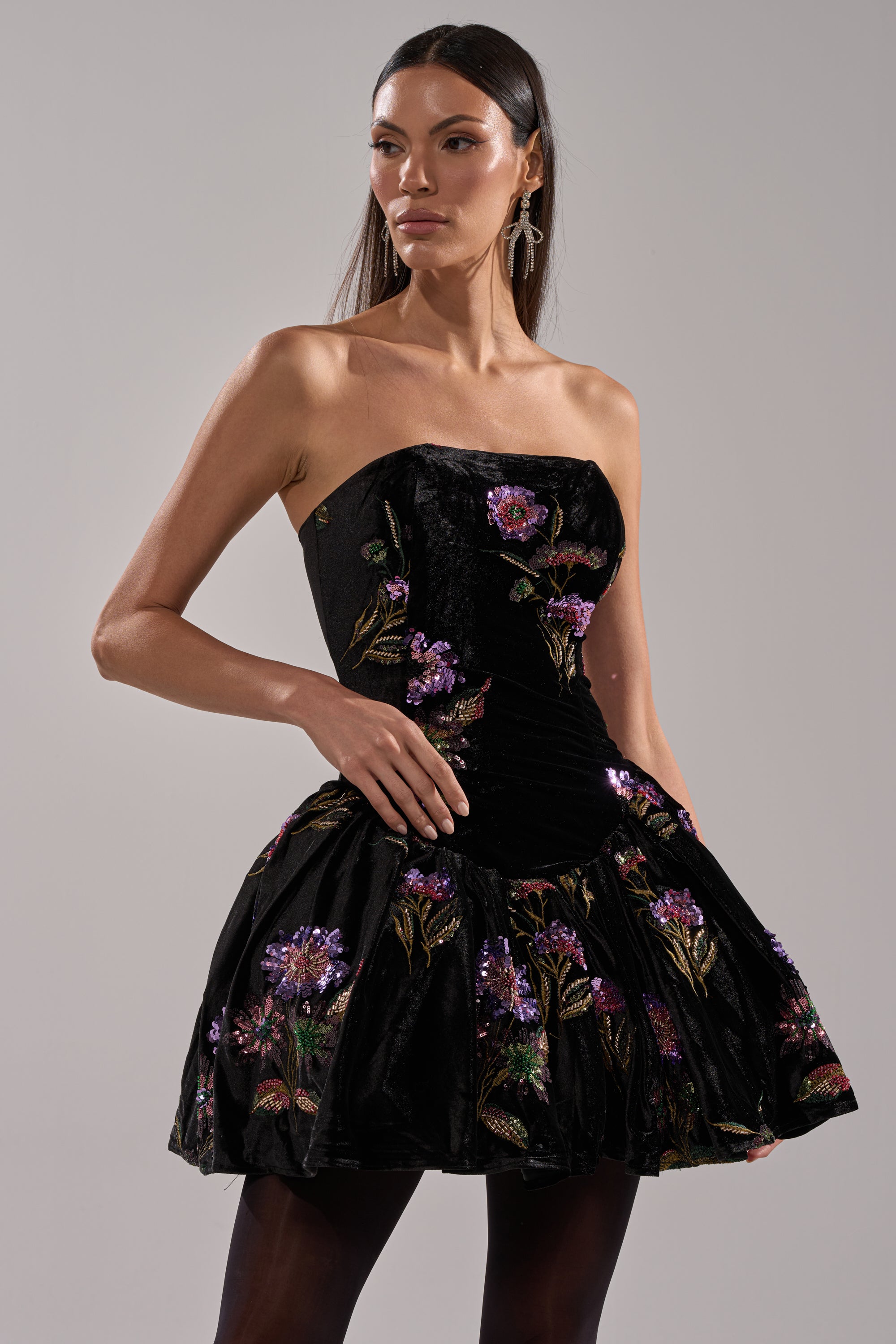 A woman poses with one hand on her hip against a light gray background, wearing the LATE NIGHT GARDEN PARTY VELVET MINI DRESS—a strapless black velvet mini dress featuring purple and gold floral embroidery.