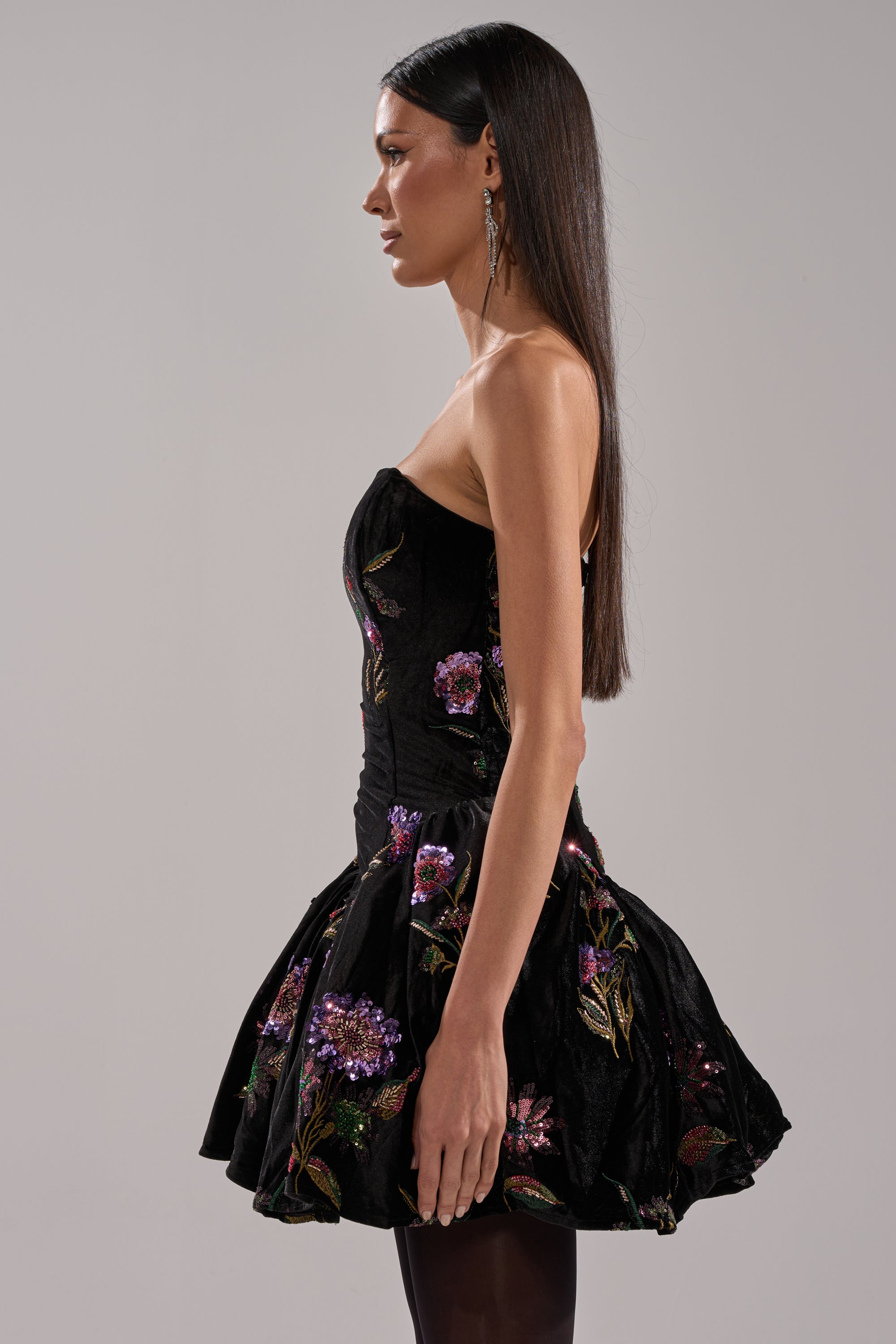 A woman stands sideways against a plain gray background, wearing the LATE NIGHT GARDEN PARTY VELVET MINI DRESS—a strapless black velvet mini dress with a short bubble skirt and colorful floral embroidery. Her long, straight hair falls down her back.