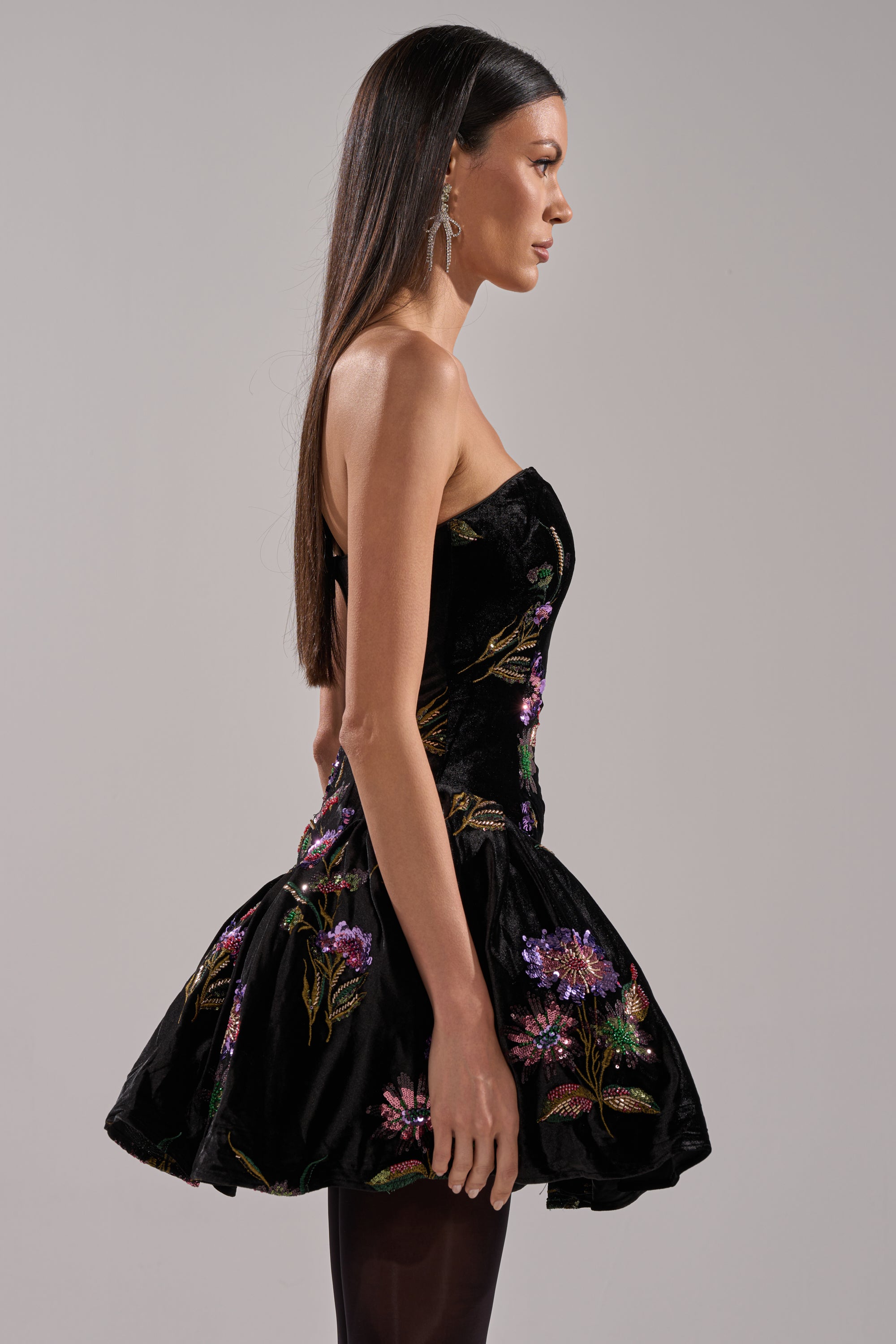 A woman with long straight brown hair models the LATE NIGHT GARDEN PARTY VELVET MINI DRESS, a strapless black mini dress with a flared skirt and colorful floral embroidery, shown in profile against a plain light gray background.