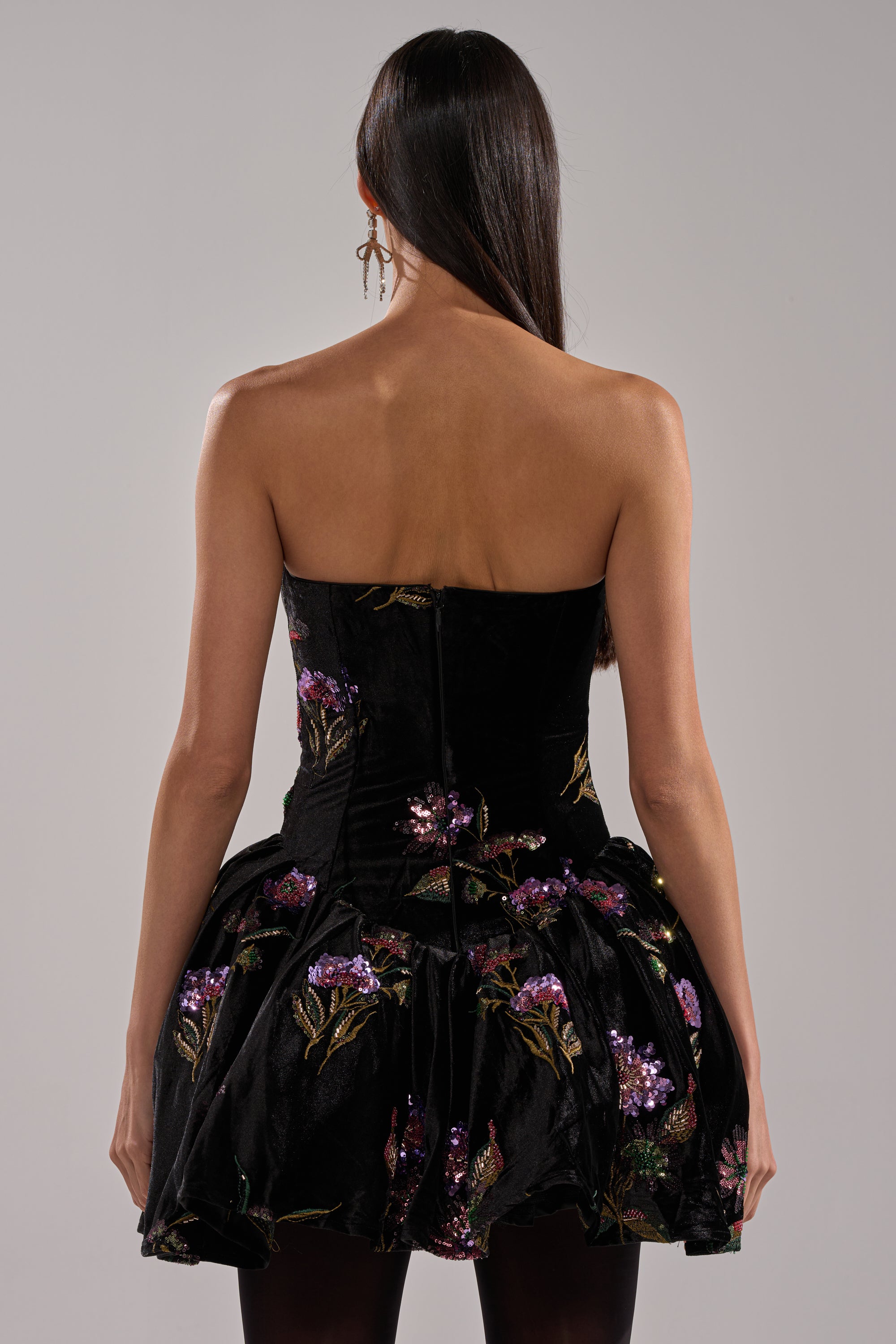 A woman with long dark hair stands with her back to the camera, wearing the LATE NIGHT GARDEN PARTY VELVET MINI DRESS—strapless, purple and gold floral embroidery, short bubble skirt—paired with earrings and dark tights.
