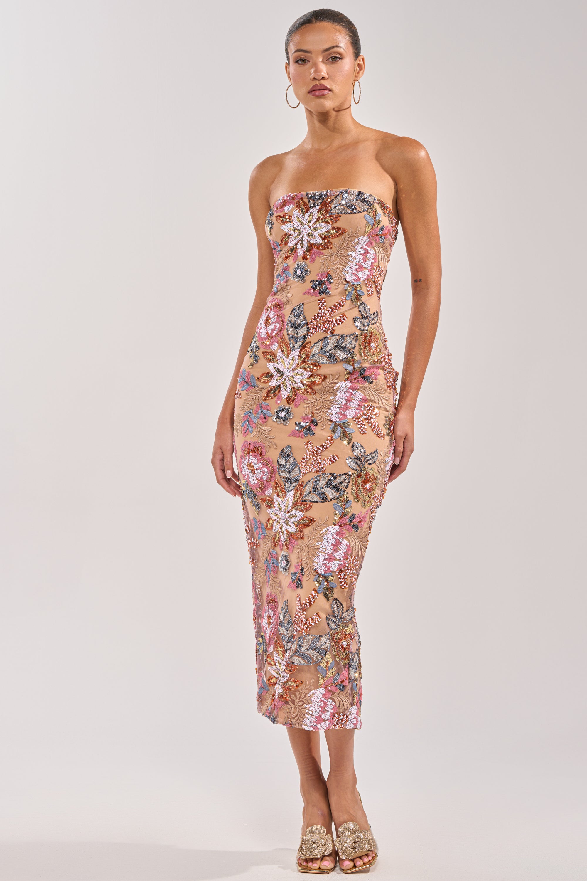 A woman models the SHOW ME LOVE EMBELLISHED MIDI DRESS, a strapless, form-fitting beige dress with colorful floral embroidery. Styled with hoop earrings and slide sandals, she poses against a light background—perfect for any occasion. 1