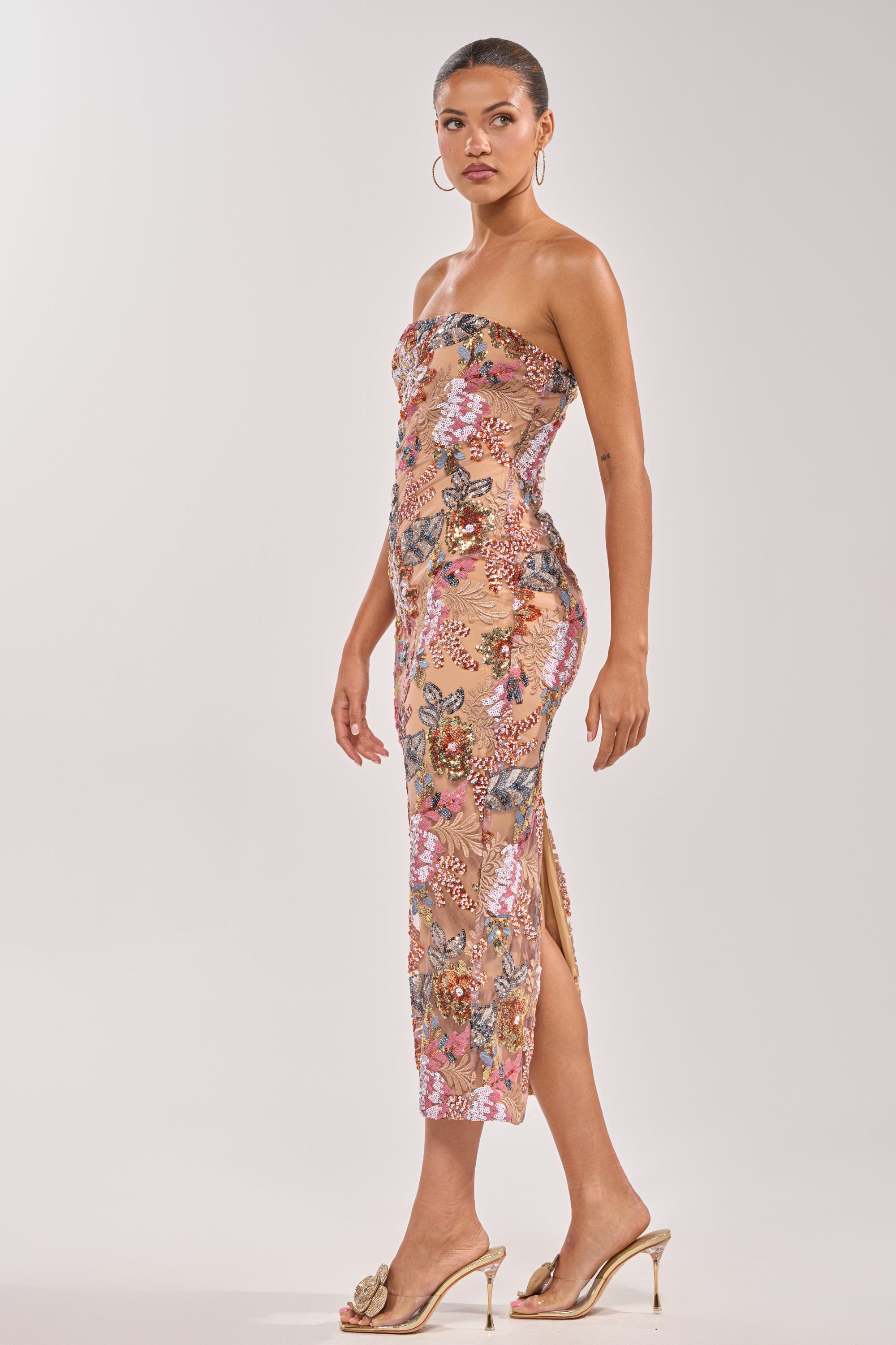 A woman poses sideways in the SHOW ME LOVE EMBELLISHED MIDI DRESS—a strapless, fitted, calf-length floral piece styled with hoop earrings, slicked-back hair, and gold high heels with bow details against a light background. 3
