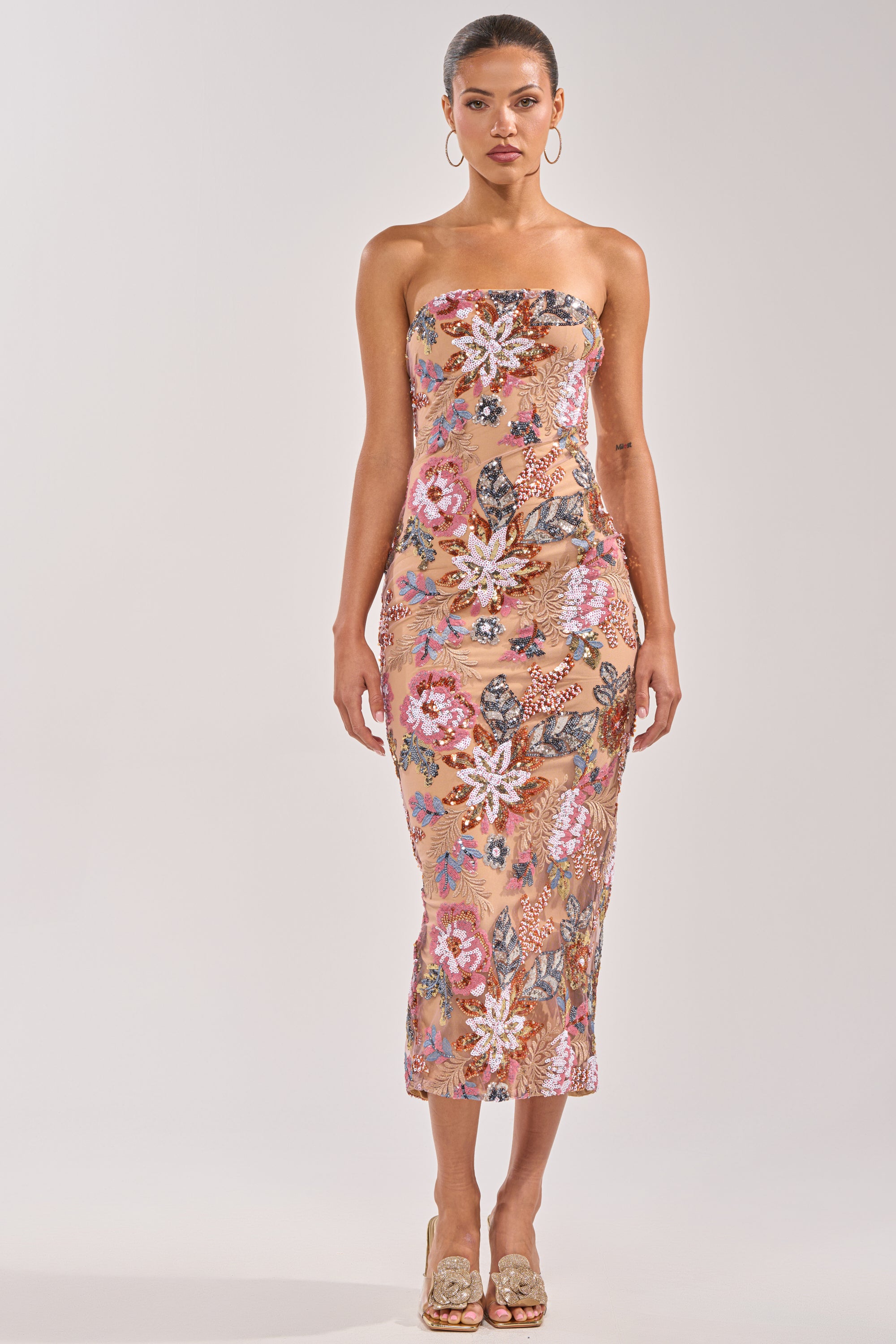 A woman faces forward wearing the SHOW ME LOVE EMBELLISHED MIDI DRESS, a strapless sequin midi with a peach base and colorful floral embroidery. She styles it with hoop earrings, heels, and tied-back hair—ideal for any occasion. 4