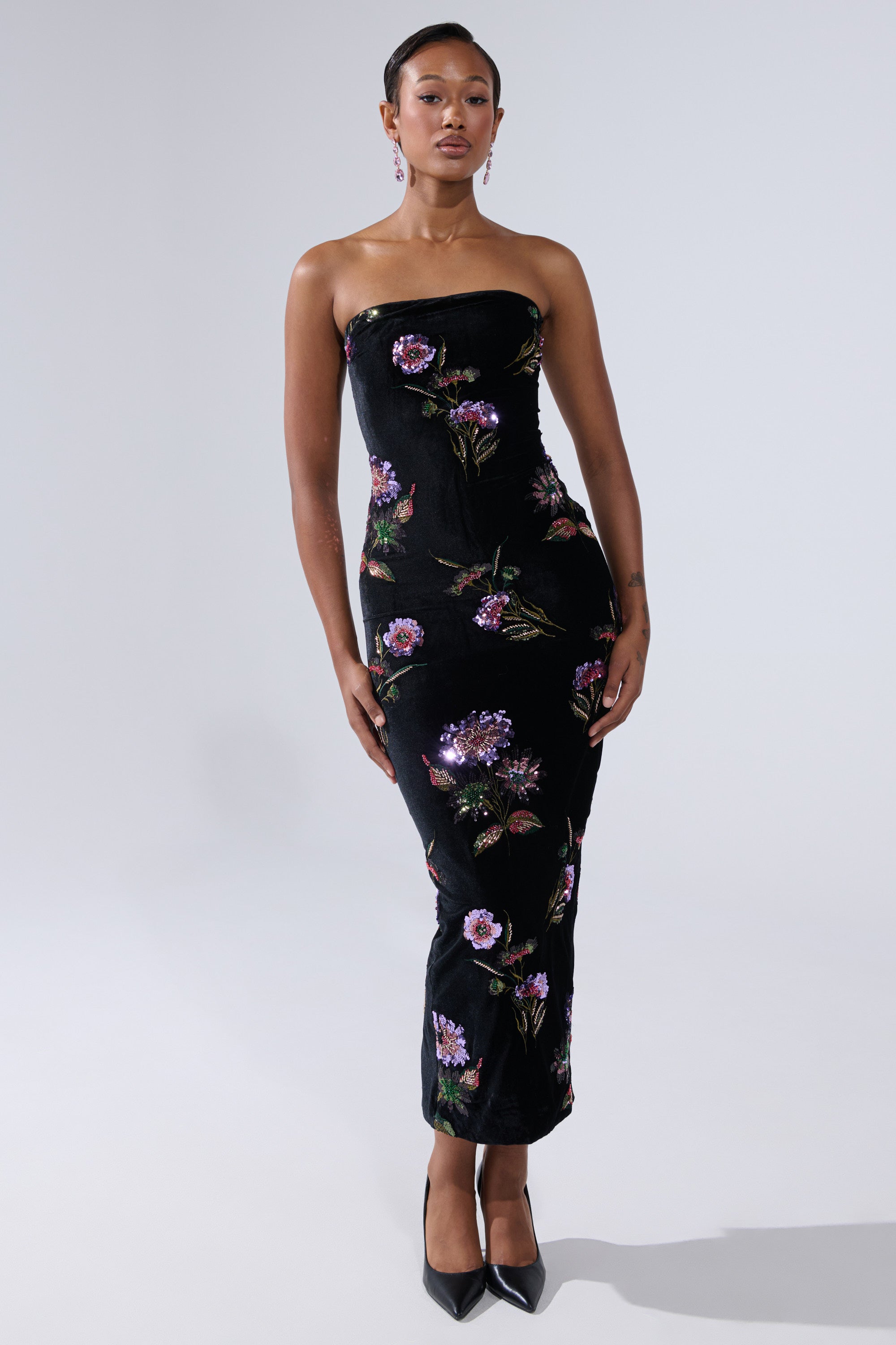 A woman wears the LUNA BEADED FLORAL VELVET MIDI DRESS, featuring colorful floral embroidery, with black pointed-toe heels and earrings. Her hair is pulled back as she poses against a plain light gray background.