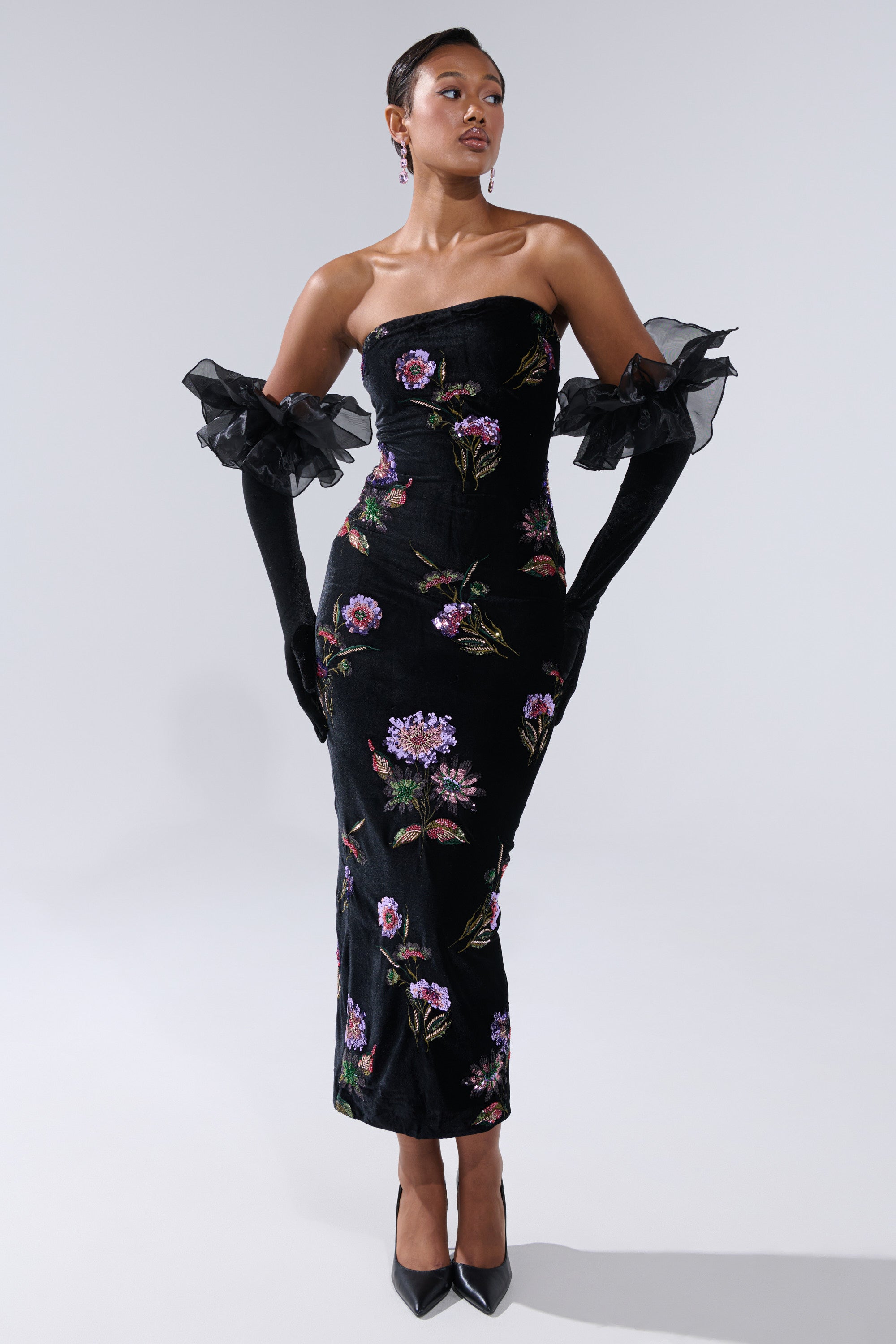 A woman wearing the LUNA BEADED FLORAL VELVET MIDI DRESS with colorful embroidery, black ruffled gloves, and black high heels stands against a plain light gray background.