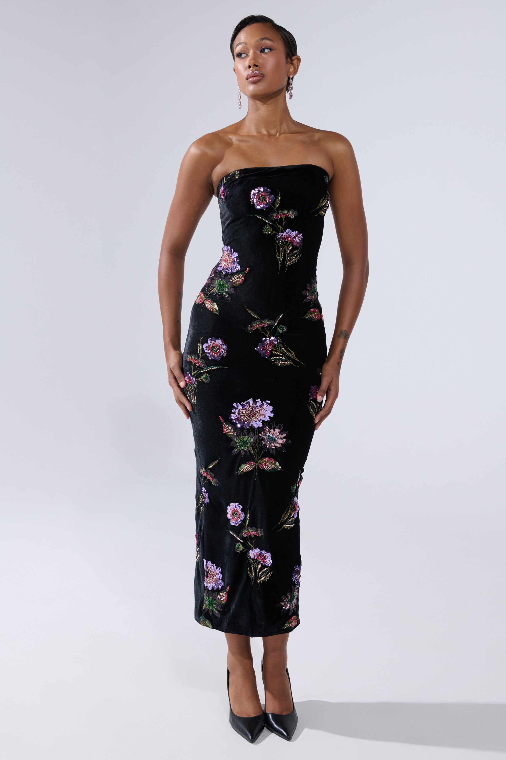 A woman wears the LUNA BEADED FLORAL VELVET MIDI DRESS, a black strapless sheath with purple floral embroidery, styled with black heels. She poses with arms relaxed by her sides against a plain light grey background.