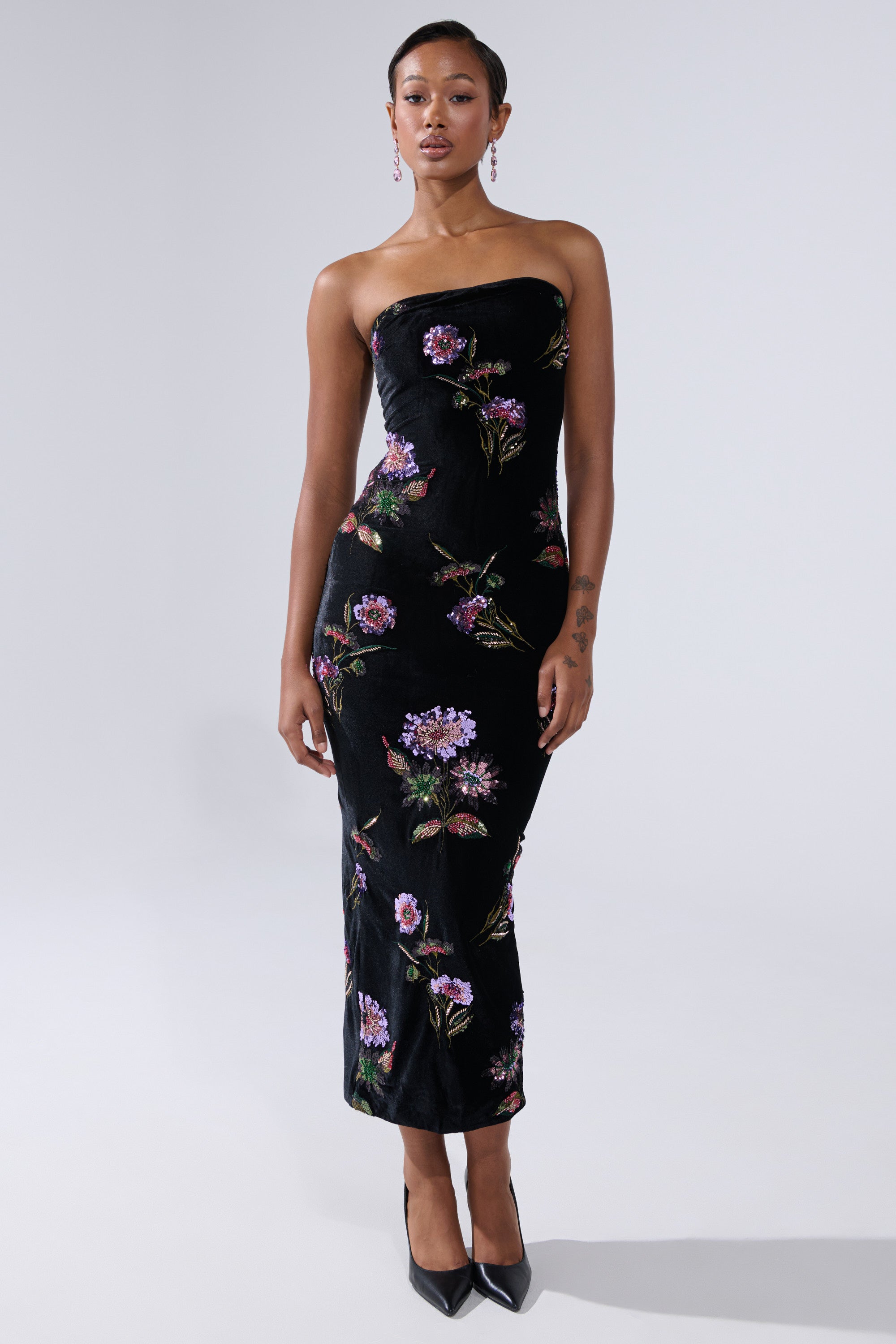 A woman wears the LUNA BEADED FLORAL VELVET MIDI DRESS—a strapless black velvet sheath with purple, pink, and green floral embroidery—paired with black pointed-toe heels, standing against a plain light gray background.