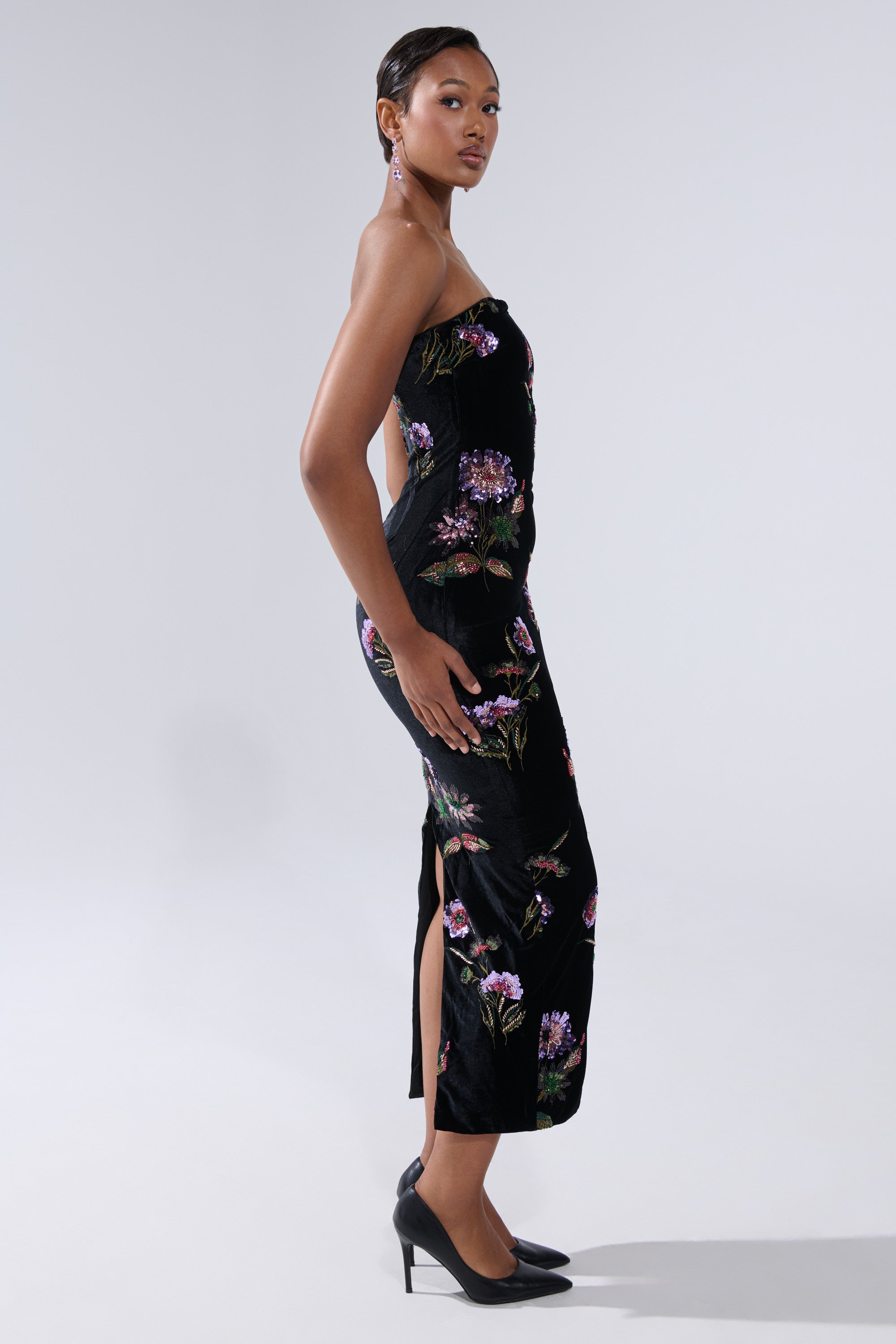 A woman poses in profile wearing the LUNA BEADED FLORAL VELVET MIDI DRESS with a floral pattern, side slit, and black high heels against a plain light gray background.