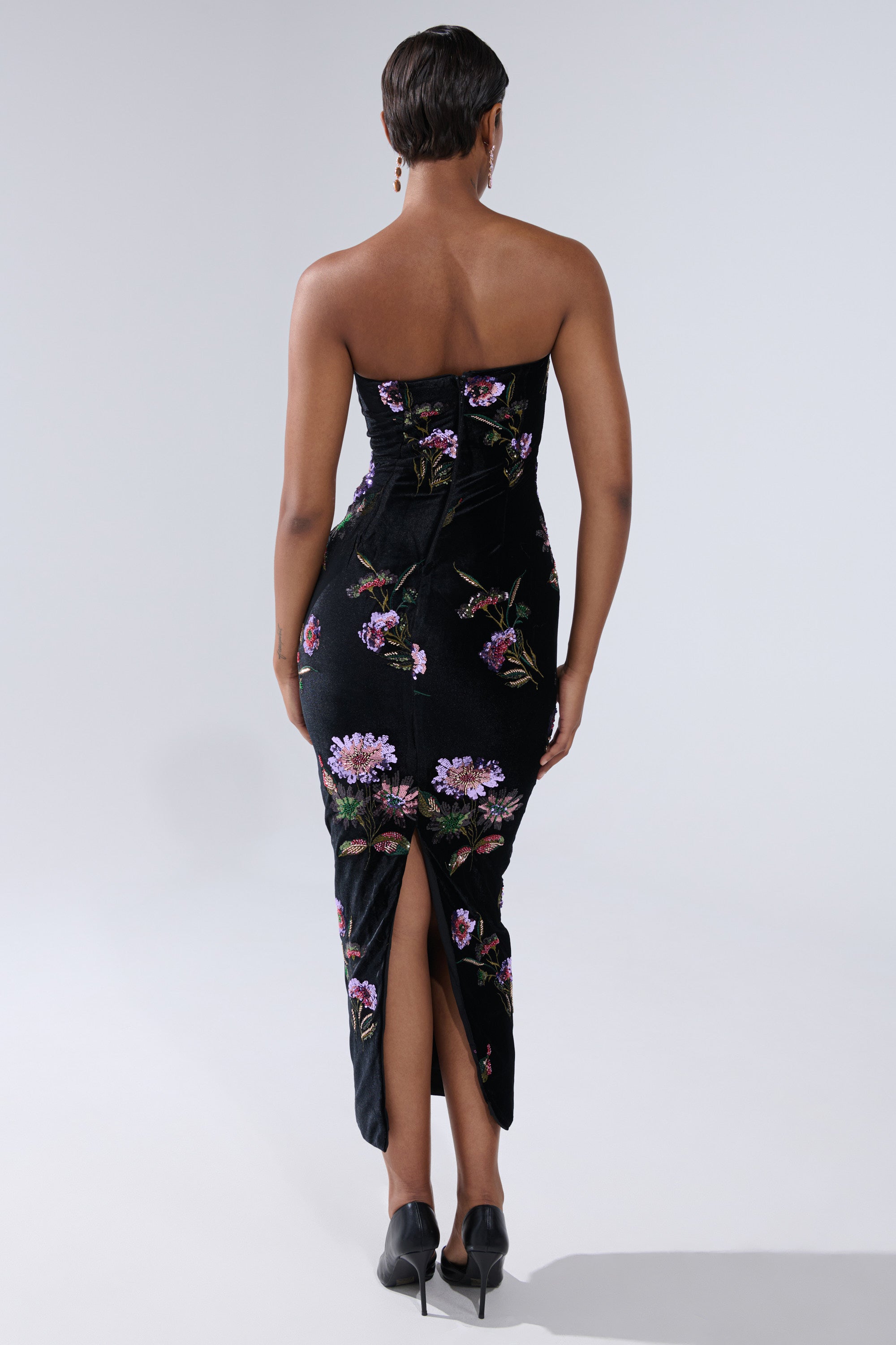 A person with short hair, seen from behind, is wearing the LUNA BEADED FLORAL VELVET MIDI DRESS—a strapless sheath dress with purple floral patterns and a back slit—paired with black heels.