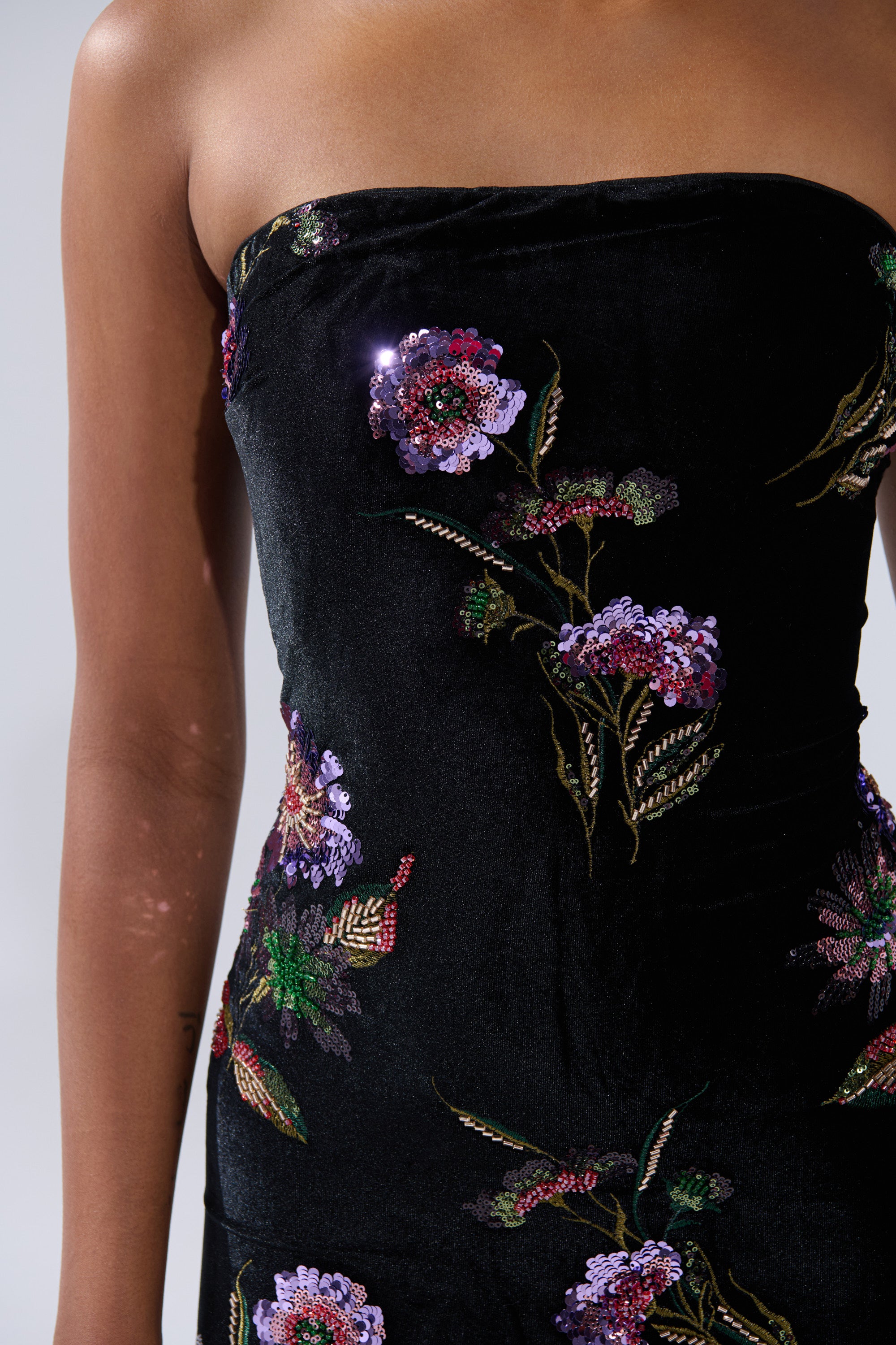 Model showcases the LUNA BEADED FLORAL VELVET MIDI DRESS, a strapless black velvet sheath featuring colorful embroidered and beaded floral designs. Only the upper body is visible against a plain background.