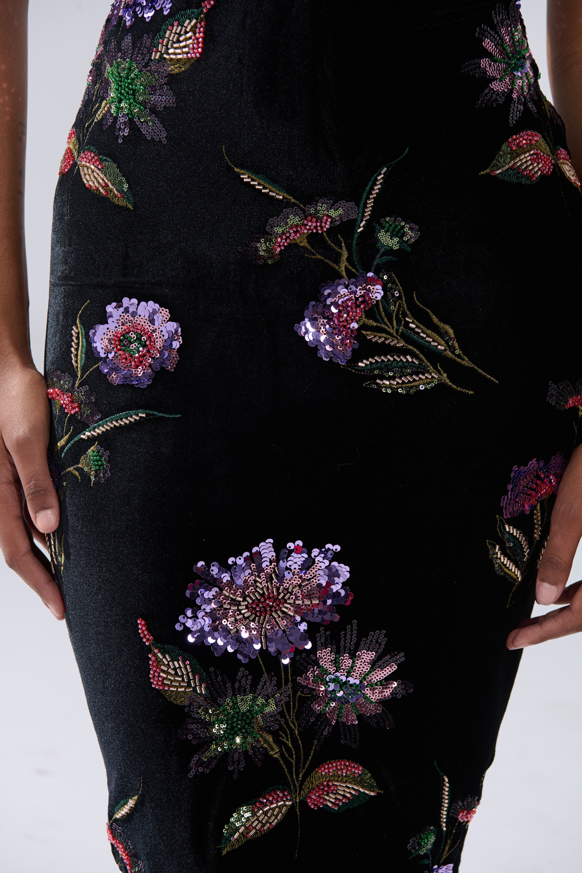 A close-up of a person wearing the LUNA BEADED FLORAL VELVET MIDI DRESS, featuring black velvet with colorful beaded floral embroidery and sequins, their hands resting by their sides.