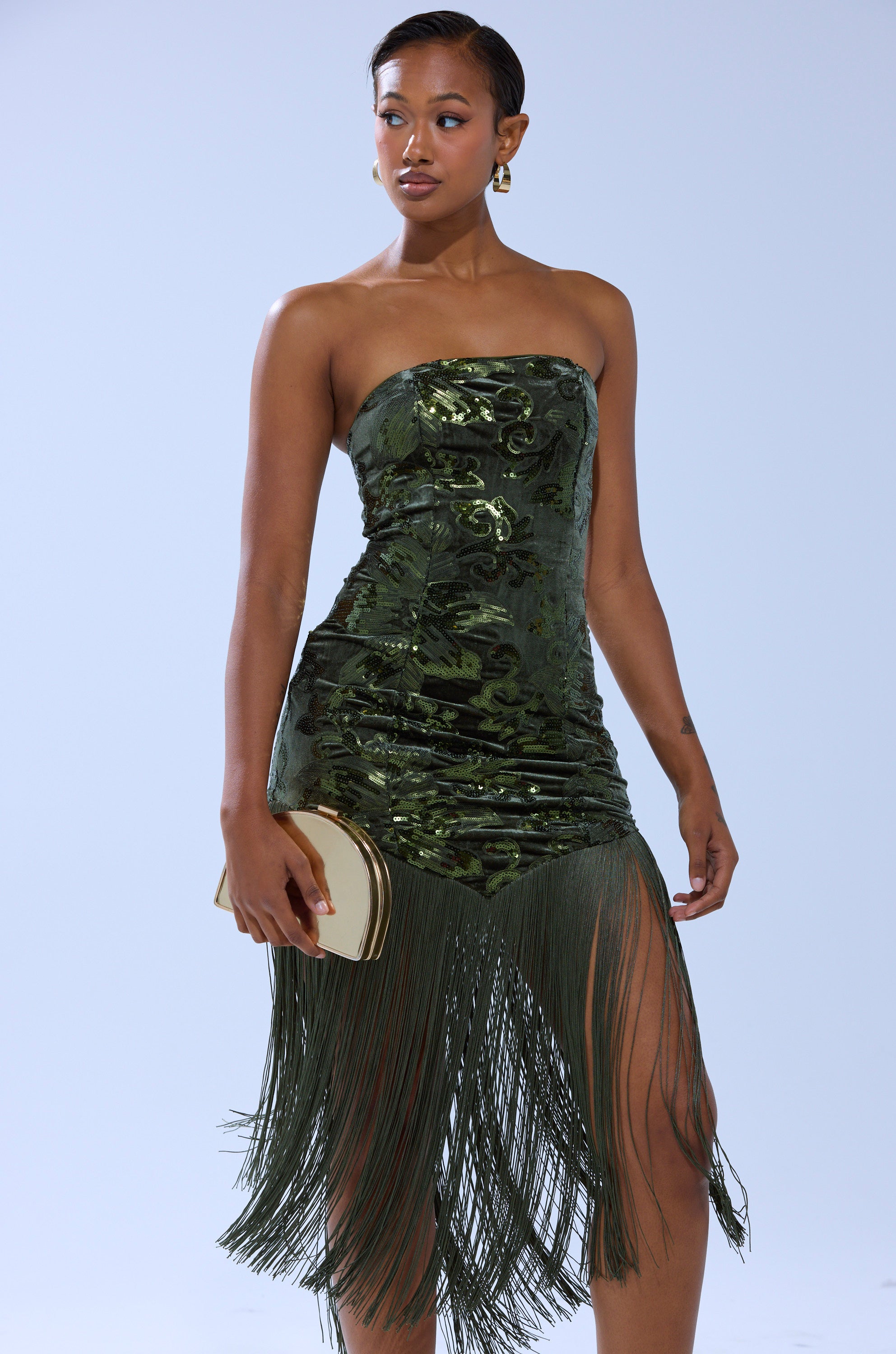 A woman in the SOFIA VELVET FRINGE PRINTED MIDI DRESS holds a gold clutch. She has short, styled hair and wears gold hoop earrings, standing against a plain light background.
