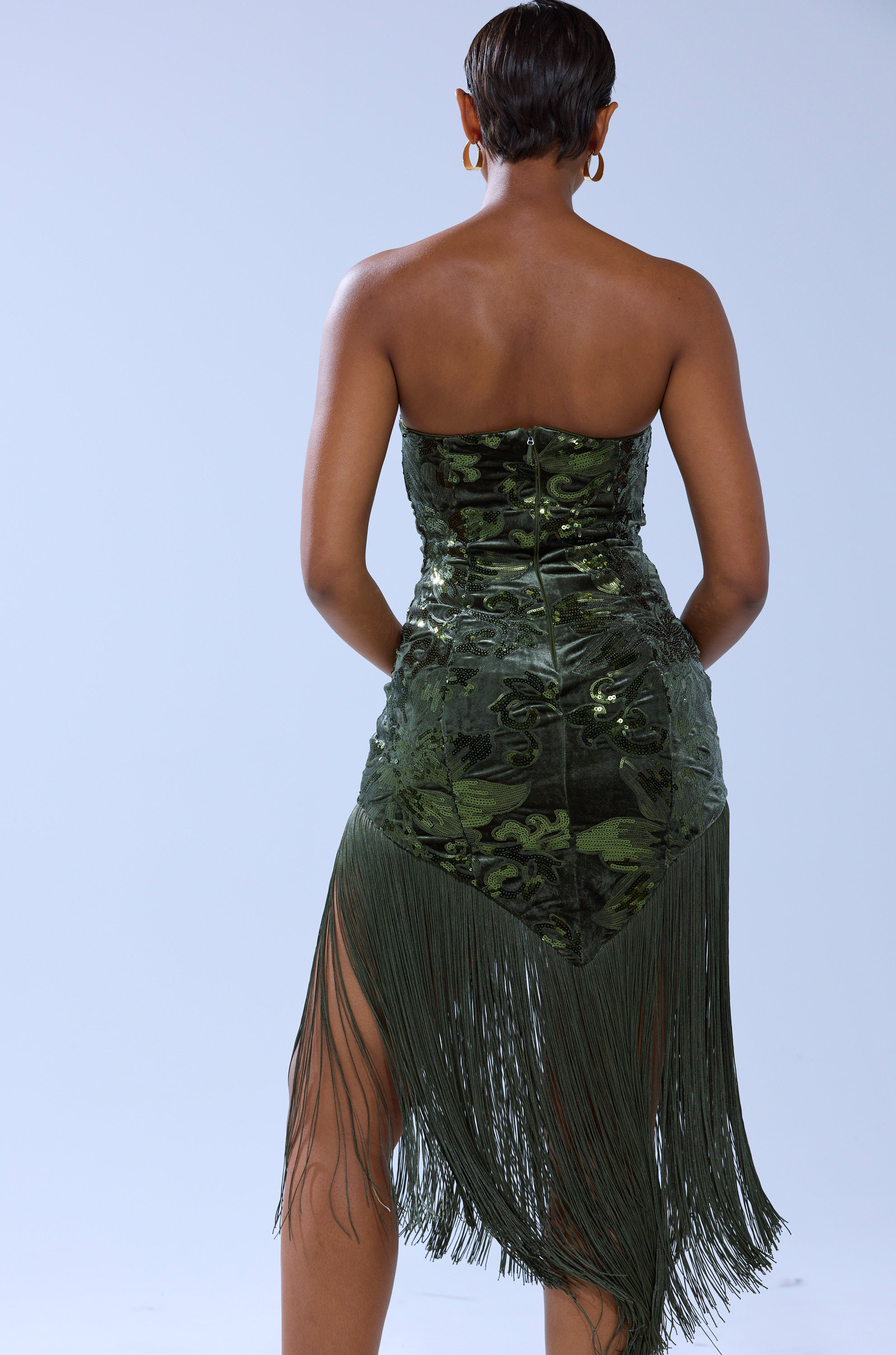 A woman with short hair faces away, wearing the SOFIA VELVET FRINGE PRINTED MIDI DRESS in dark green and gold hoop earrings, standing against a plain light background.