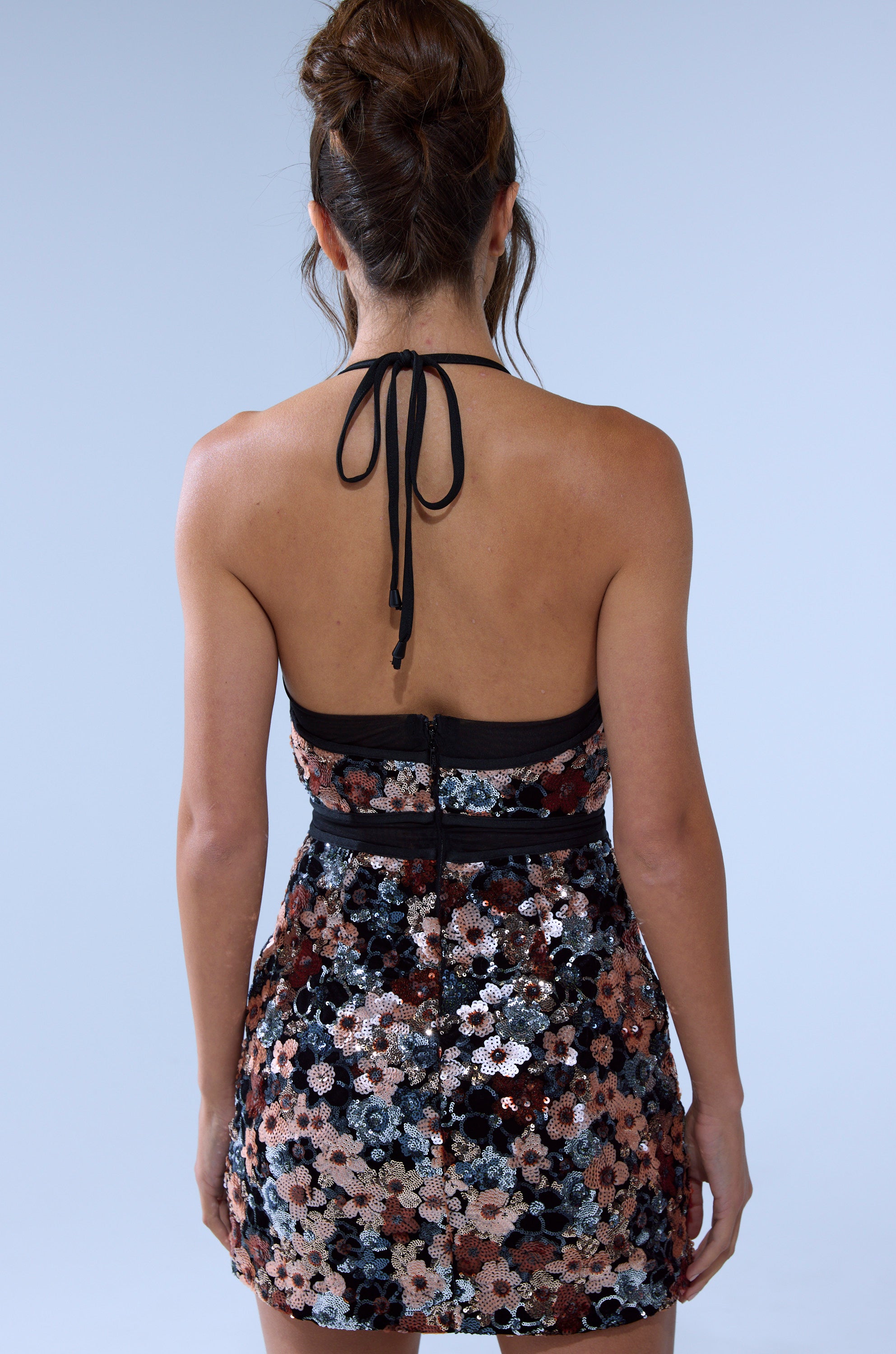 A woman with her hair in a loose bun faces away, wearing the ALONNA SEQUIN MINI DRESS—a halter mini with a colorful floral sequin pattern and open back—against a plain light gray background.