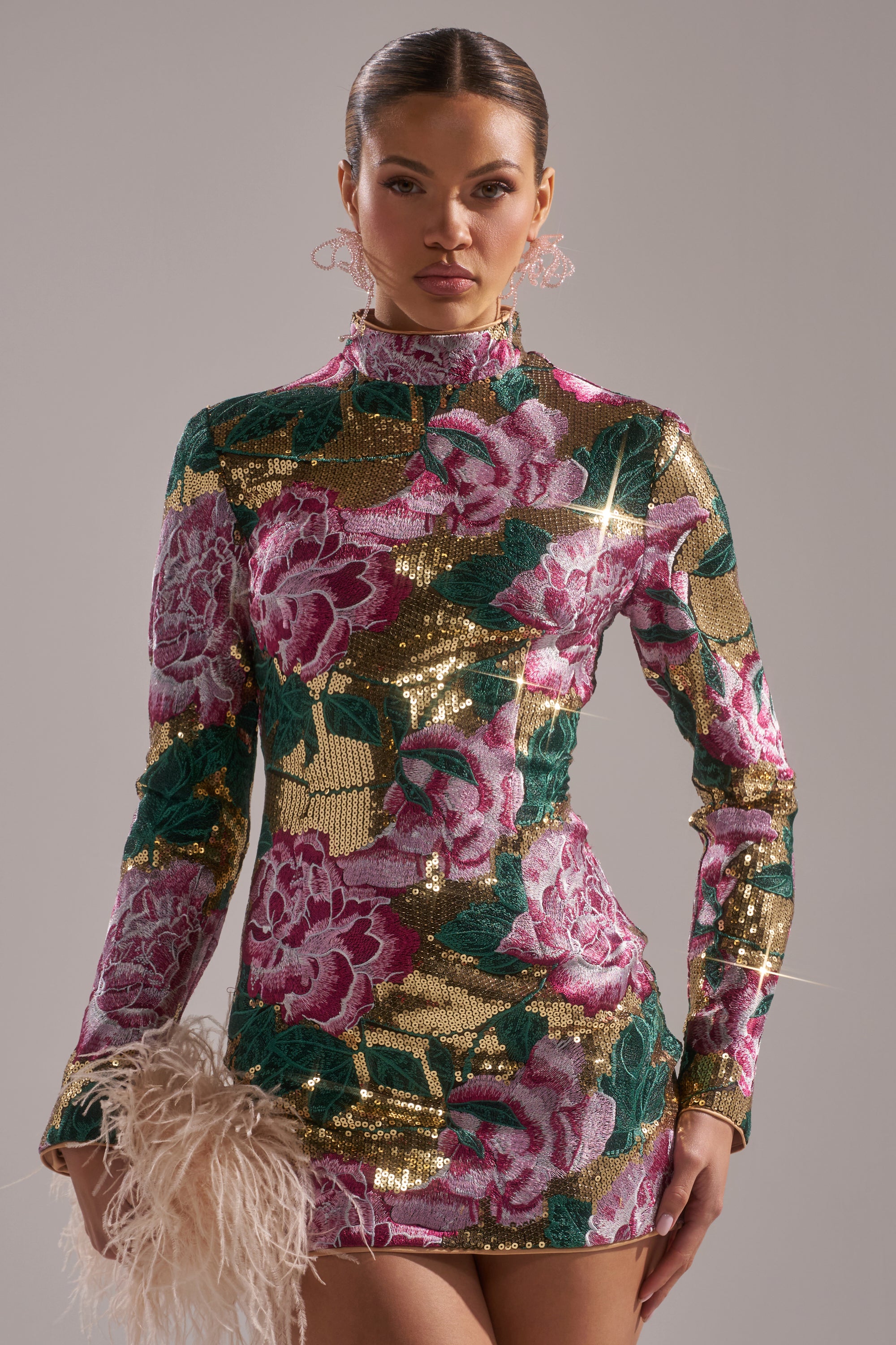 A woman wears the FLORAL BABY SEQUIN MINI DRESS with long sleeves and vibrant pink, green, and gold patterns, posing with a feathered accessory against a plain background.