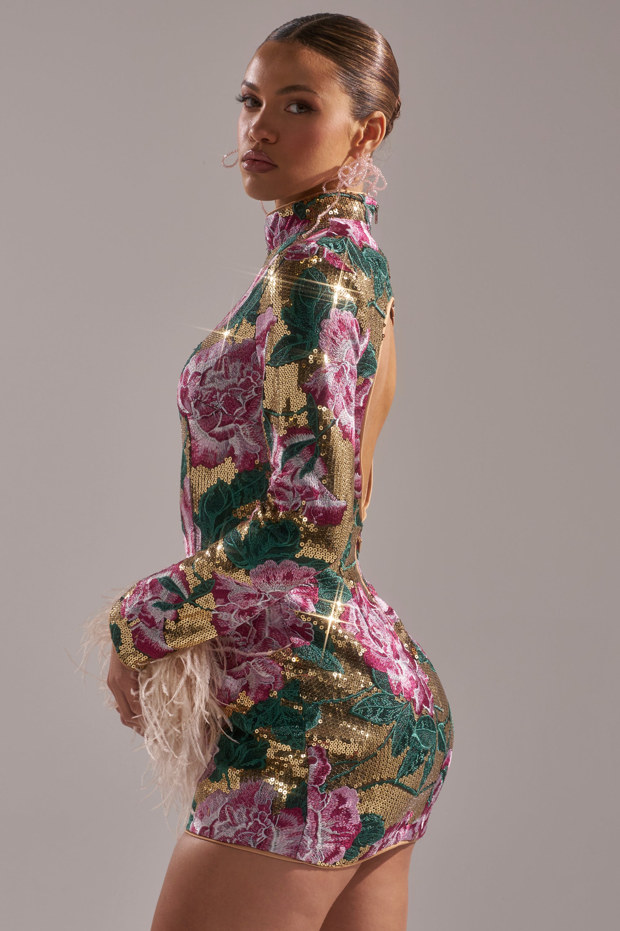 A woman poses in the FLORAL BABY SEQUIN MINI DRESS, a form-fitting mini with a high neck, feathered cuffs, open back, and pink, green, and gold floral sequins shimmering against a plain light background.