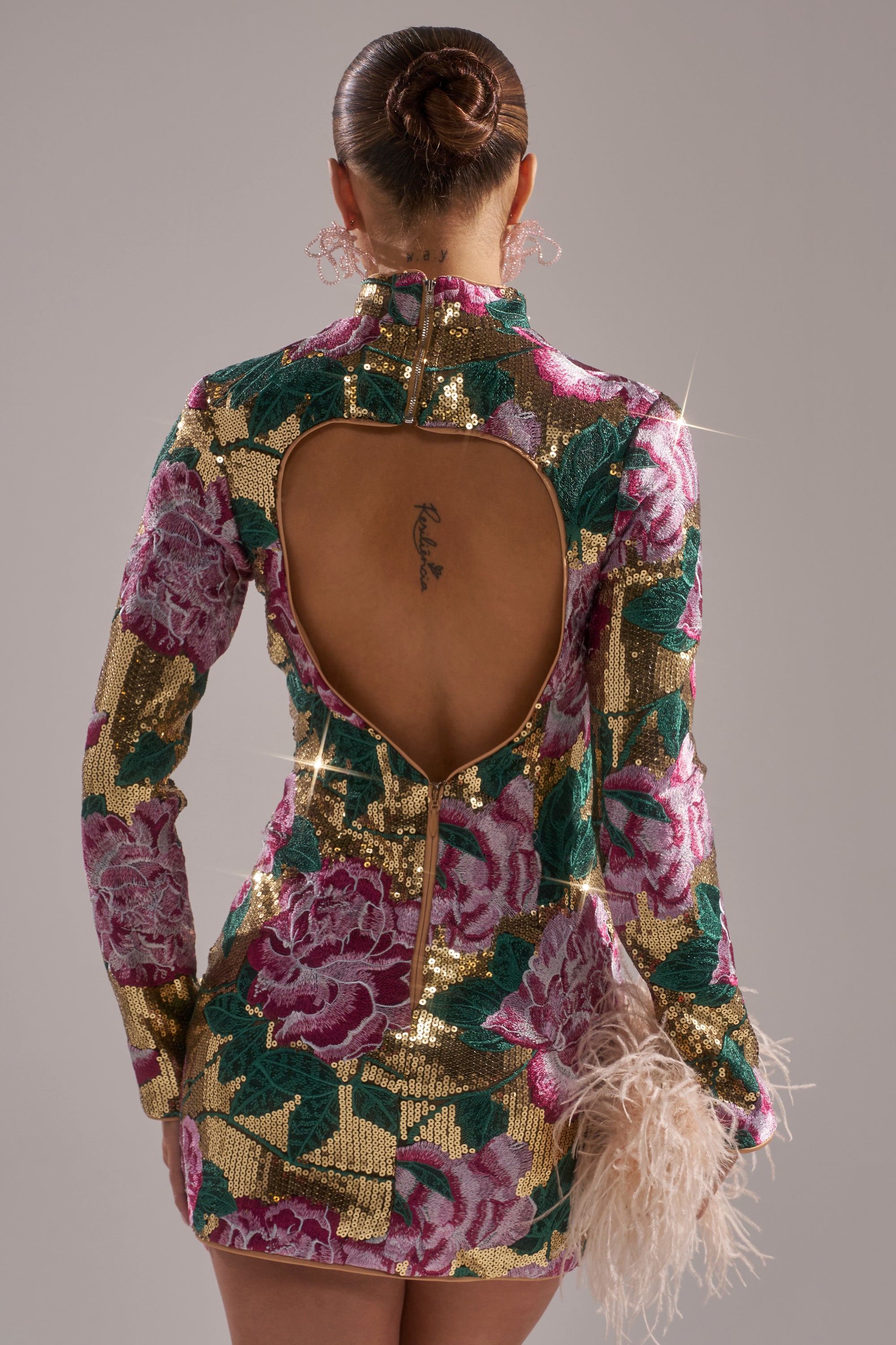 A person with a sleek bun wears the FLORAL BABY SEQUIN MINI DRESS featuring an open back that reveals a small tattoo. They hold a feathered accessory while standing against a plain background.