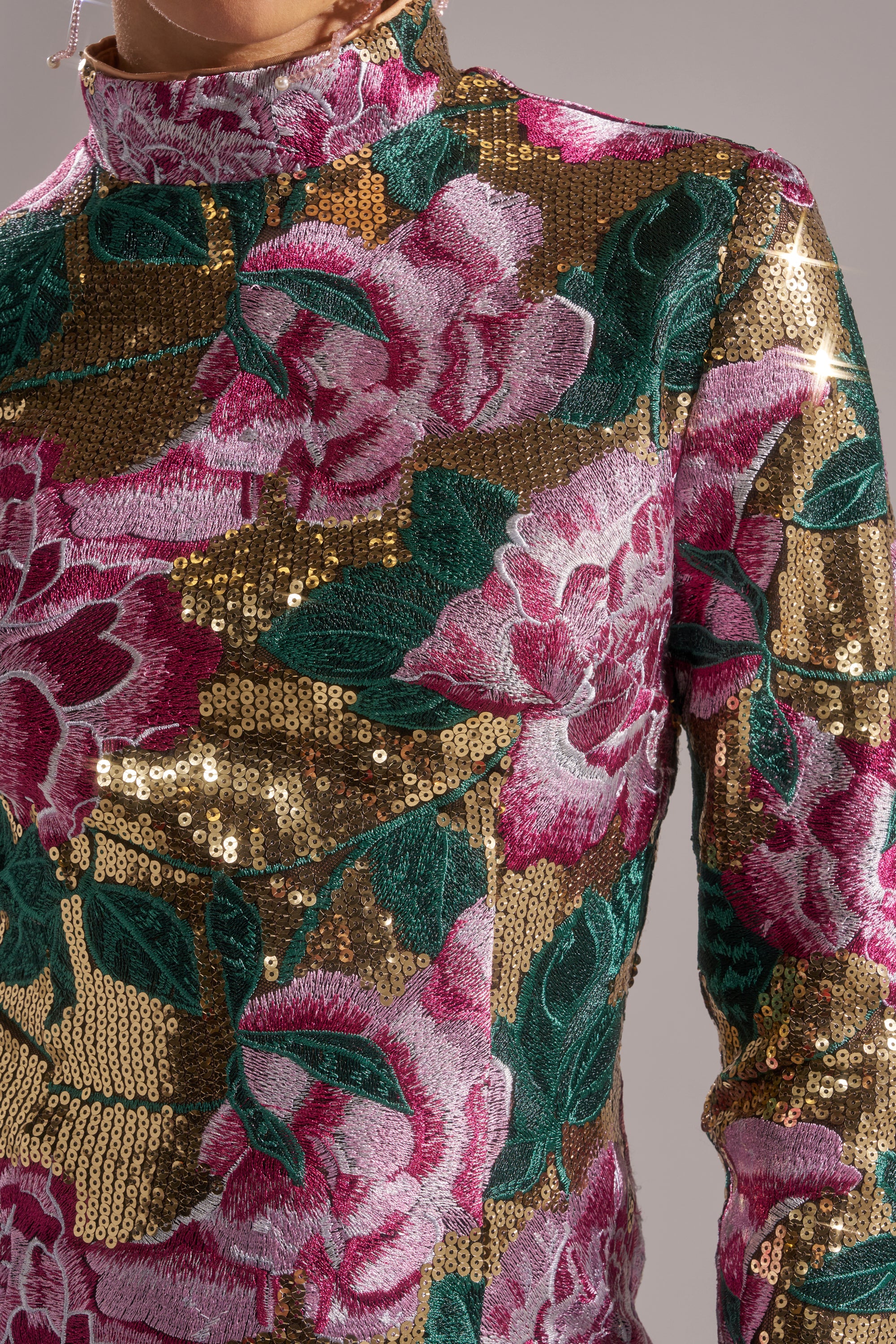 Close-up of a person wearing the FLORAL BABY SEQUIN MINI DRESS—a high-neck, long-sleeved holiday party dress shimmering with gold sequins, embroidered pink flowers, and green leaves that beautifully catch the light.