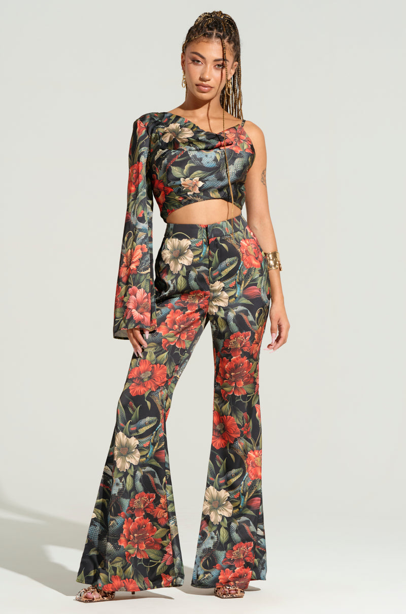 A woman stands confidently in a fashionable outfit featuring a one-shoulder asymmetrical crop top and the DON'T GET IT TWISTED PRINTED SATIN TROUSER from the AKIRA Label. The ensemble is adorned with a vibrant tropical floral print in shades of red, green, and cream. She wears hoop earrings and a gold bracelet.