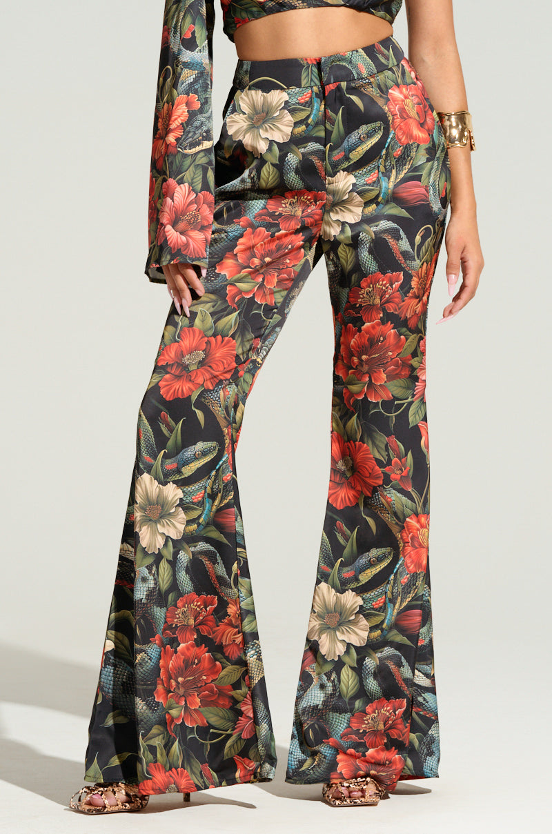 A person wearing the vibrant "DON'T GET IT TWISTED PRINTED SATIN TROUSER," adorned with bold red, white, and green tropical floral prints that flare at the bottom. Part of a matching floral-patterned sleeve and a gold bracelet are also visible against a plain background.