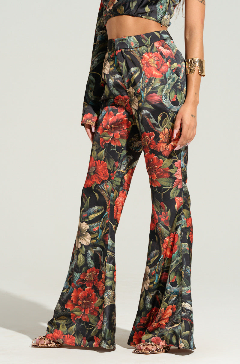 A person is wearing a coordinated outfit from the AKIRA Label. The outfit features the DON'T GET IT TWISTED PRINTED SATIN TROUSER, which is high-waisted with wide legs, showcasing a tropical floral print and snake pattern. The long-sleeved crop top matches perfectly with its bright red flowers and green foliage on a black background, and the look is complemented by a gold cuff bracelet.