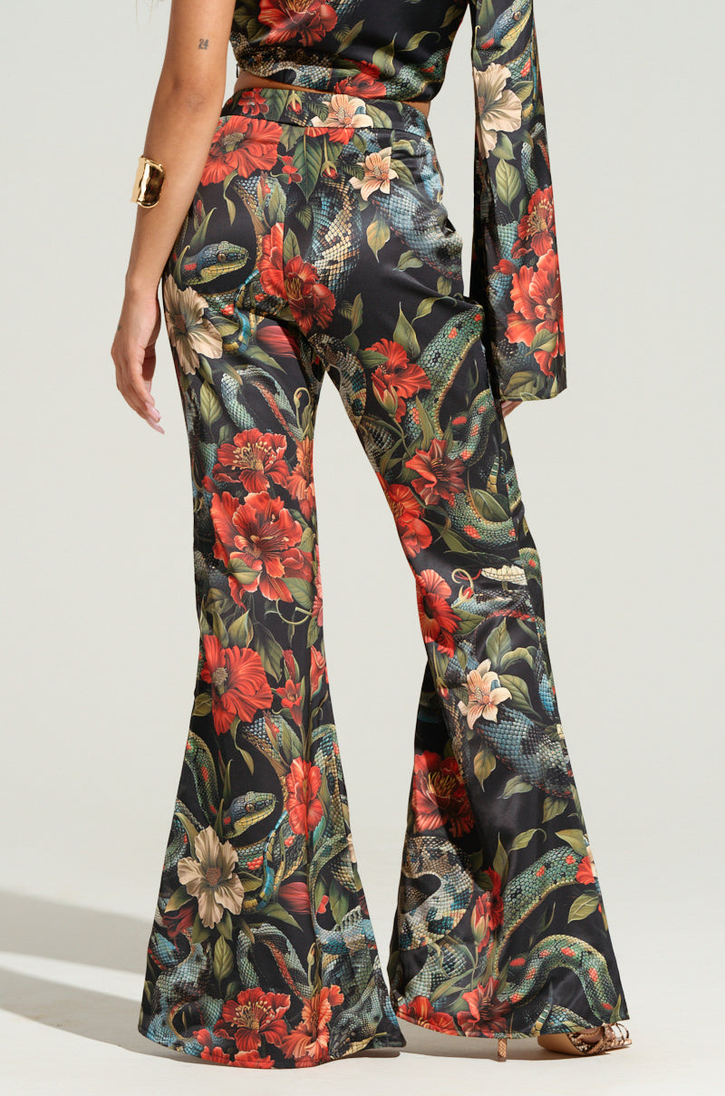 A person wearing the DON'T GET IT TWISTED PRINTED SATIN TROUSER from the AKIRA Label, featuring high-waisted, flared pants with a vibrant, tropical floral print. The ensemble includes a matching sleeve draped from the shoulder. The plain, light background highlights the colorful details of this printed satin trouser outfit.