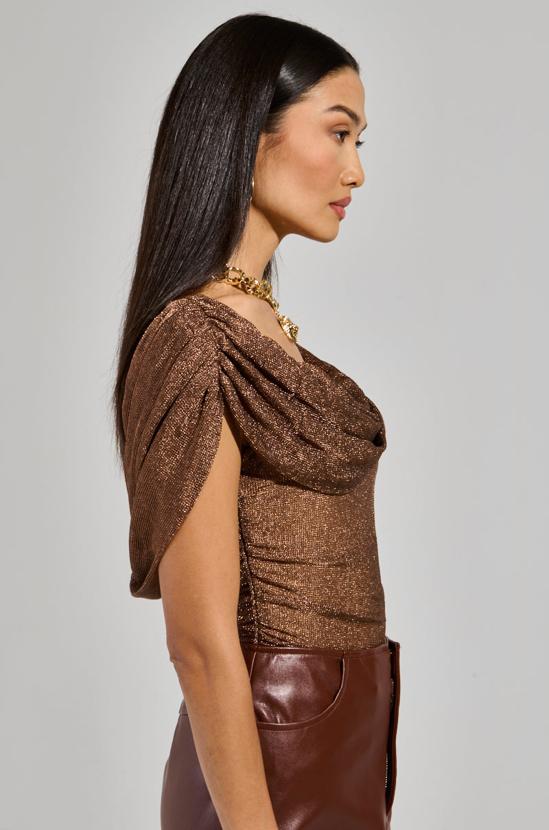 Profile of a woman with long dark hair, wearing the OUT OF HIS LEAGUE OFF THE SHOULDER TOP in shimmering brown with draped sleeves, paired with a high-waisted leather skirt. She's accessorized with a gold necklace and hoop earrings, set against a plain background.