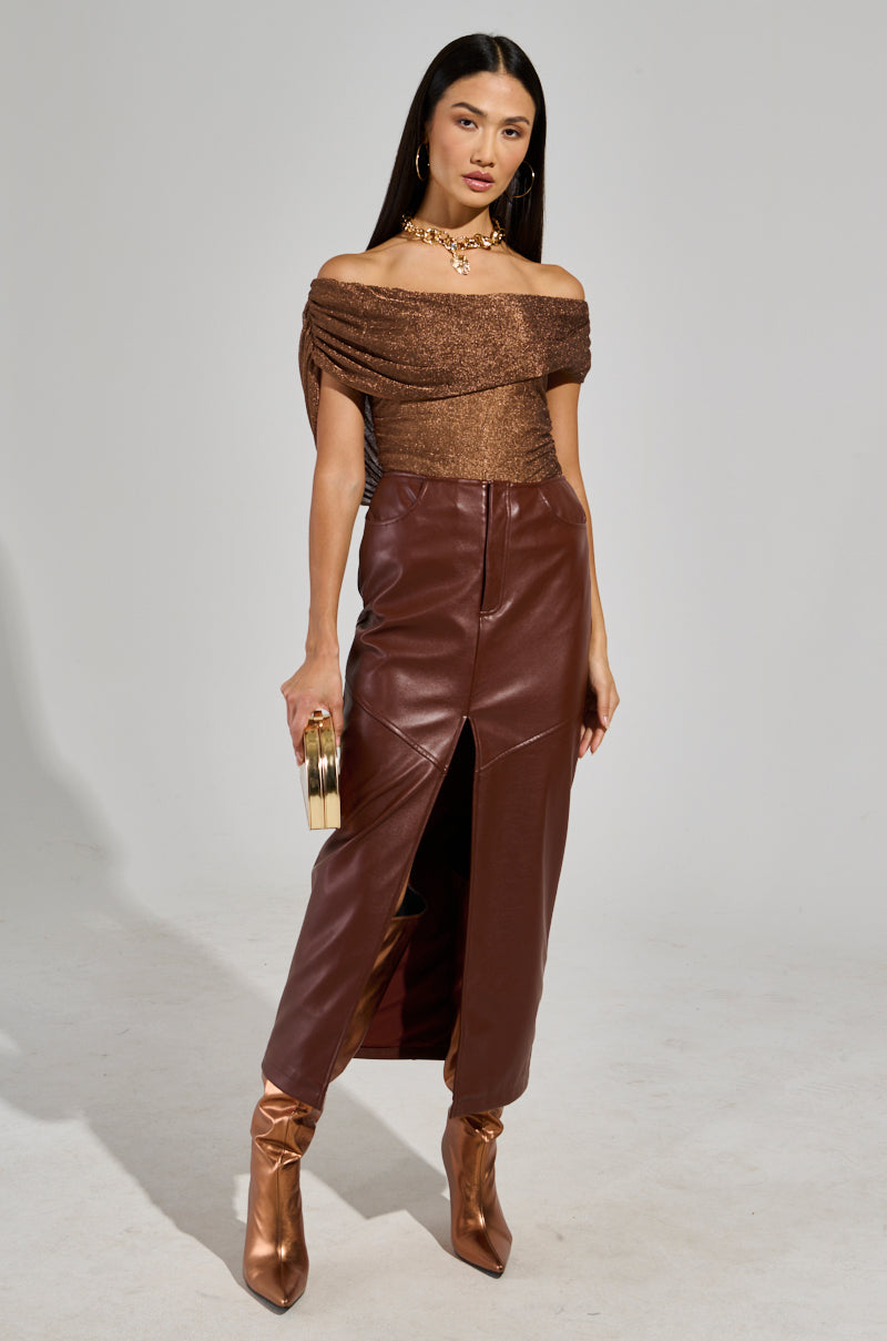 A woman poses in a chic ensemble, featuring the "OUT OF HIS LEAGUE OFF THE SHOULDER TOP" with a metallic mesh knit design. She pairs it with a brown leather skirt that has ruched construction and a front slit, complemented by bronze ankle boots. Her look is accessorized with hoop earrings, a gold necklace, and she holds a small handbag against a plain background.