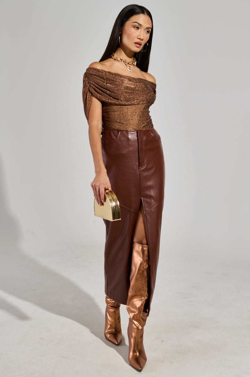 A woman wearing the "OUT OF HIS LEAGUE OFF THE SHOULDER TOP" and a long brown leather skirt with a front slit poses against a plain background. She holds a gold clutch and wears metallic knee-high boots, along with a shimmering gold necklace.
