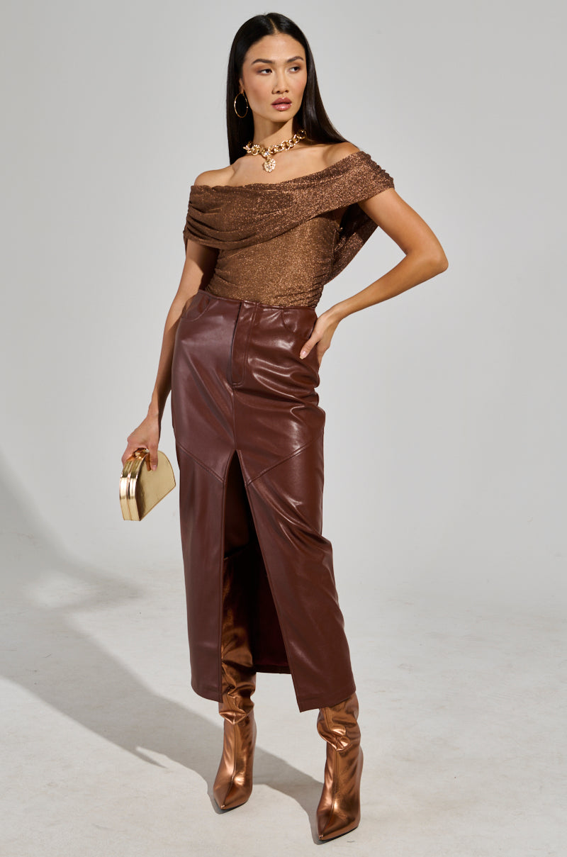 A woman poses in the sleek "OUT OF HIS LEAGUE OFF THE SHOULDER TOP" with a stylish ruched design. She pairs it with a long brown leather skirt featuring a front slit, matching brown boots, and carries a small gold handbag. Her chunky gold necklace complements her long, straight hair flowing elegantly down.