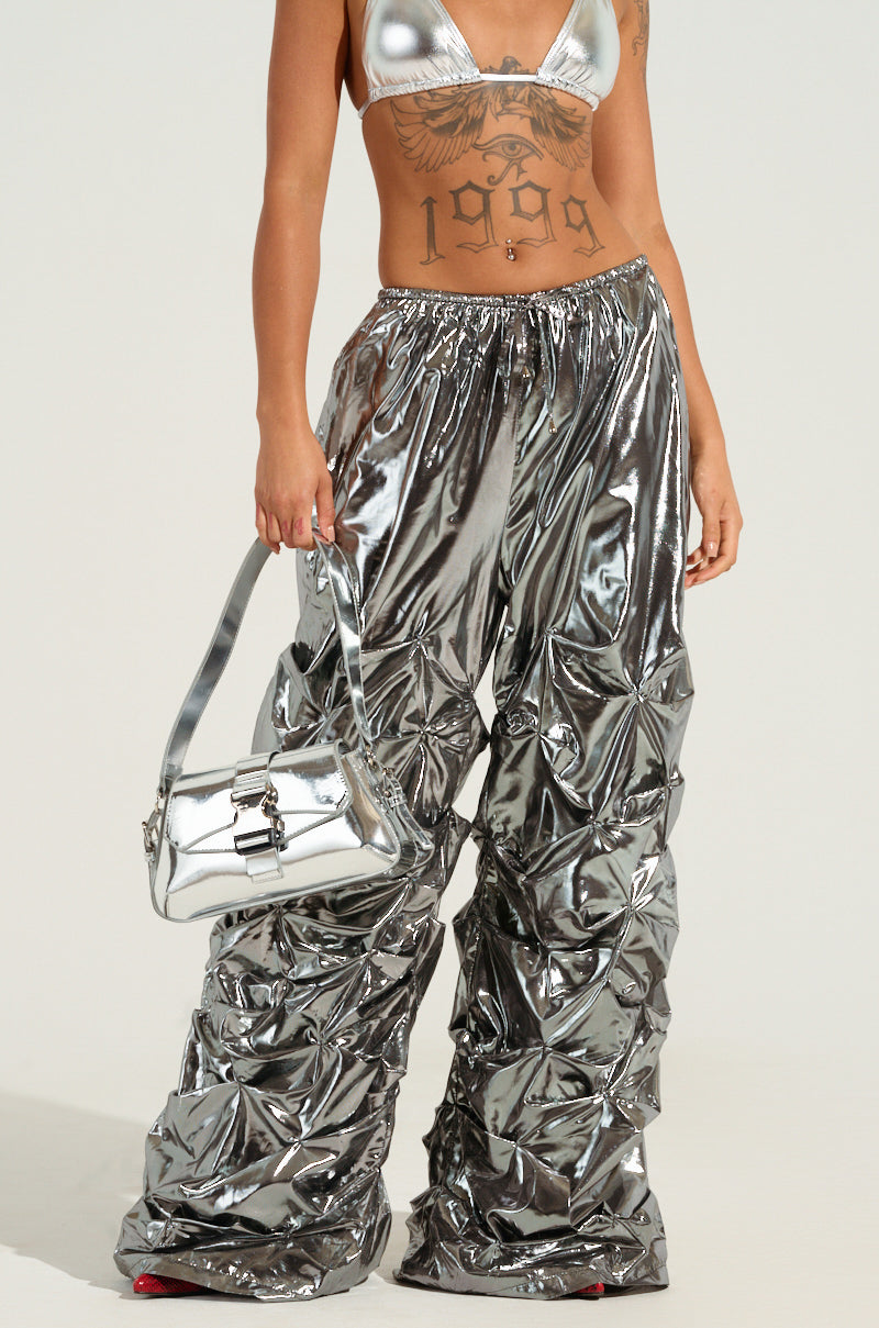 A person wearing an oversized metallic crop top and COINCIDENTAL LOVE OVERSIZED METALLIC RUCHED PANT with a wide leg silhouette holds a matching silver handbag. The reflective, bold outfit is styled with intricate tattoos on their torso, capturing the essence of coincidental love.