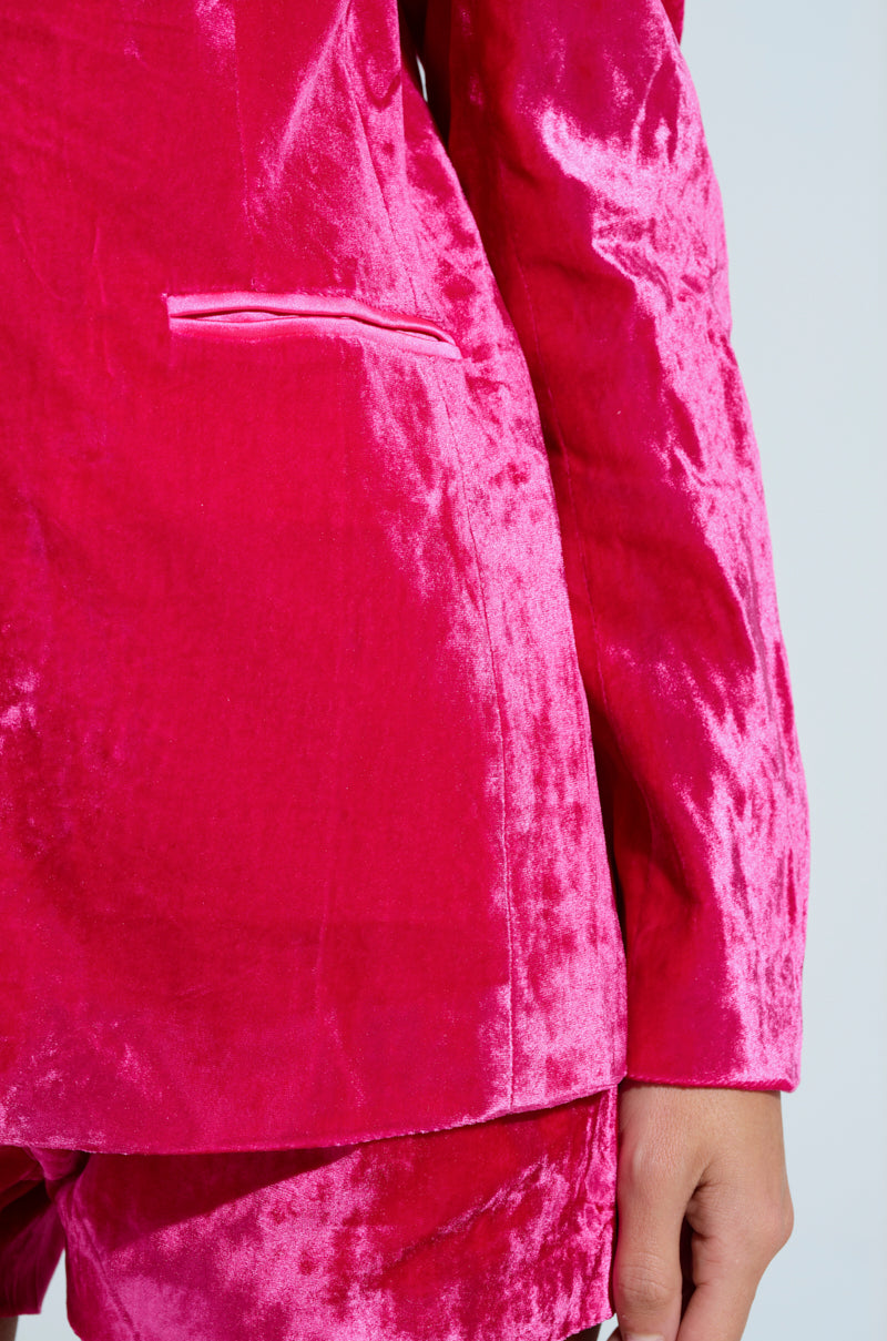 A person models the Pretty in Pink Velvet Blazer and matching shorts, showcasing a bright pink suit with a smooth, shiny texture, satin-lined interior, and visible jacket pocket. The plain, light-colored background highlights the vibrant outfit.