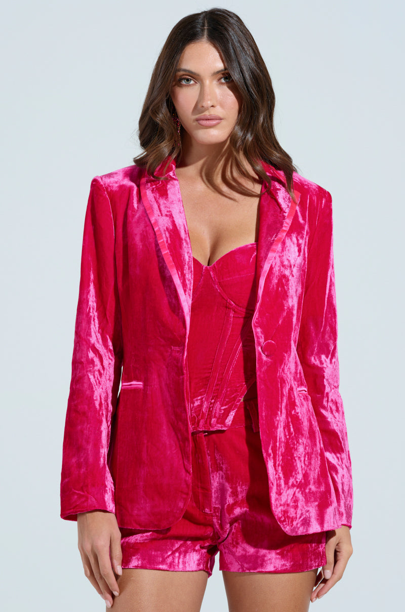 A woman, wearing the PRETTY IN PINK VELVET BLAZER with a satin-lined interior, pairs it with a corset and shorts. Her long, wavy hair flows down as she stands against a plain light background, gazing directly at the camera.