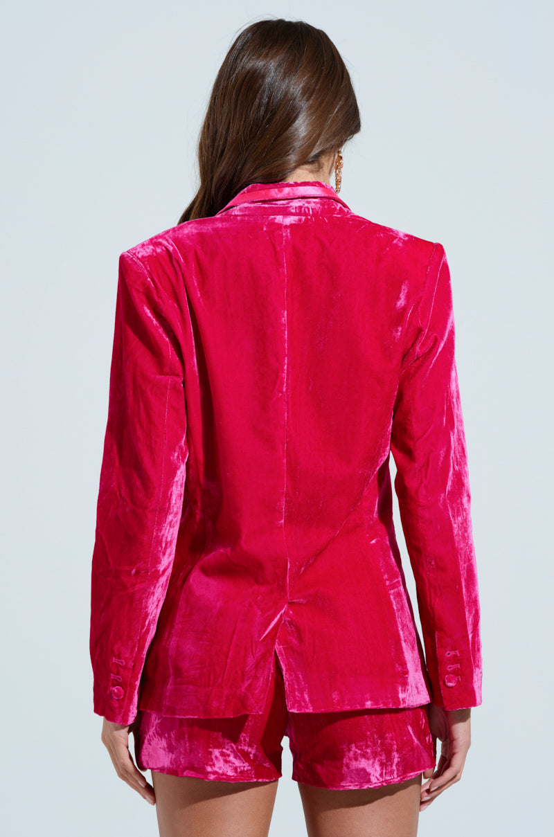 A person with long hair is turned away from the camera, wearing a PRETTY IN PINK VELVET BLAZER and eye-catching matching shorts against a light background.