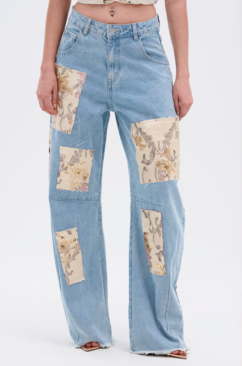 A person wears ROYAL ENERGY BROCADE PATCH RELAXED DENIM—light-wash jeans featuring large rectangular beige brocade patches on the front, raw hems, and paired with a cropped top.