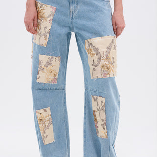 A person wears ROYAL ENERGY BROCADE PATCH RELAXED DENIM—light-wash jeans featuring large rectangular beige brocade patches on the front, raw hems, and paired with a cropped top.