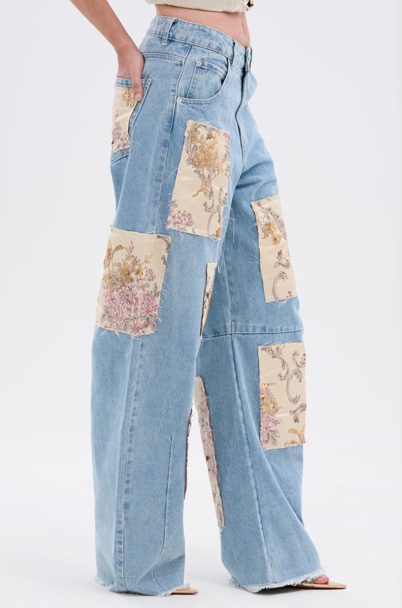 A person wearing ROYAL ENERGY BROCADE PATCH RELAXED DENIM featuring light-wash, wide-leg jeans with large cream brocade patches on the front and sides. Only the lower torso and legs are shown against a plain background.