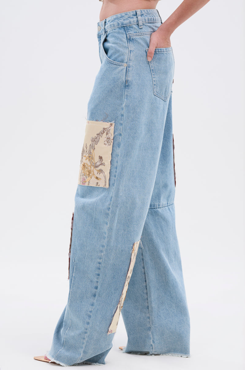 A person stands against a plain white background wearing ROYAL ENERGY BROCADE PATCH RELAXED DENIM—light-wash, wide-leg jeans with raw hems and beige floral brocade patches on the thigh and lower leg.