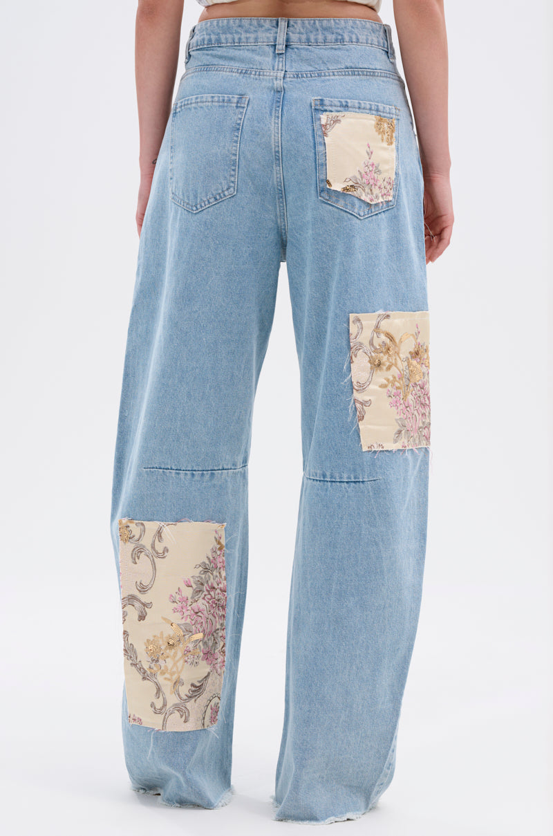 A person wearing ROYAL ENERGY BROCADE PATCH RELAXED DENIM—light-wash, wide-leg jeans featuring bold brocade patches on the back pocket, left thigh, and right calf—stands against a white background.