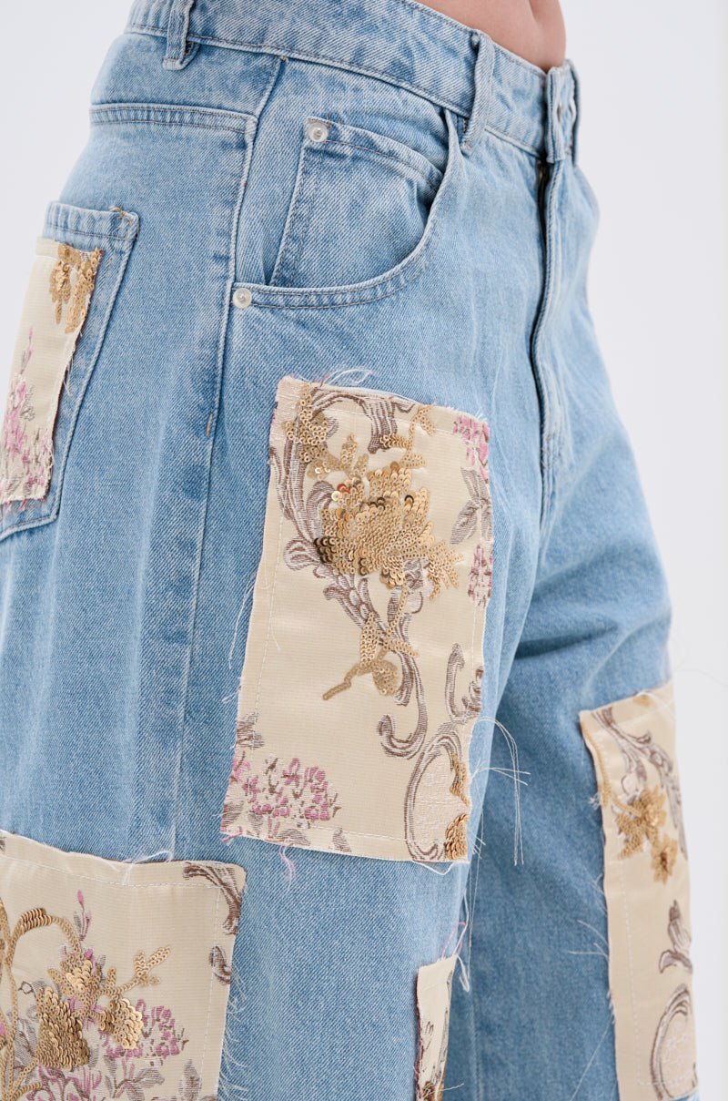 Close-up of ROYAL ENERGY BROCADE PATCH RELAXED DENIM, light-wash jeans with large cream, gold, and pink brocade patches on legs and pockets; frayed edges add a vintage, handmade touch.